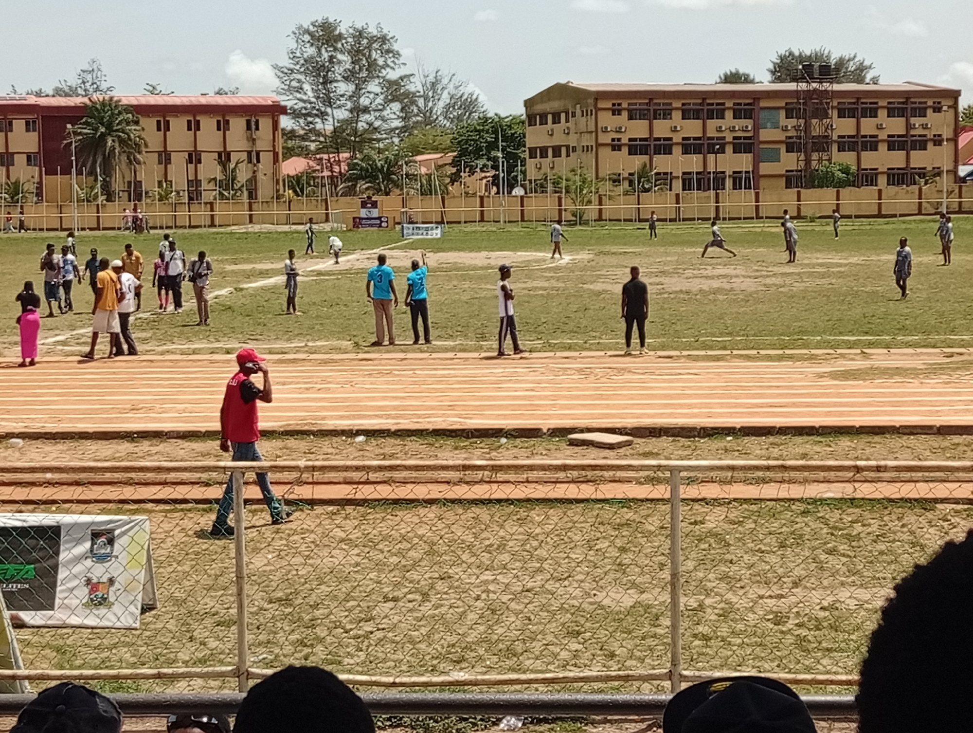 The image shows a cricket match in progress on a grassy field. In the background, there are two large, yellow buildings with multiple windows, likely educational institutions or dormitories. A running track surrounds the cricket field, with several lanes visible. Several people are scattered across the field, some actively participating in the cricket match while others appear to be spectators or officials. One man in a red shirt and jeans is walking along the track, seemingly talking on a cell phone. A chain-link fence separates the field from the seating area where the photo was taken. The lower part of the image reveals that the view is from a spectator area, with the heads of several people in the foreground. A sign with logos is partially visible behind the fence. The sky is overcast, and the light suggests it's daytime. This location may be in Lagos, Nigeria, judging by the style of the buildings and the presence of a local flag on the sign.