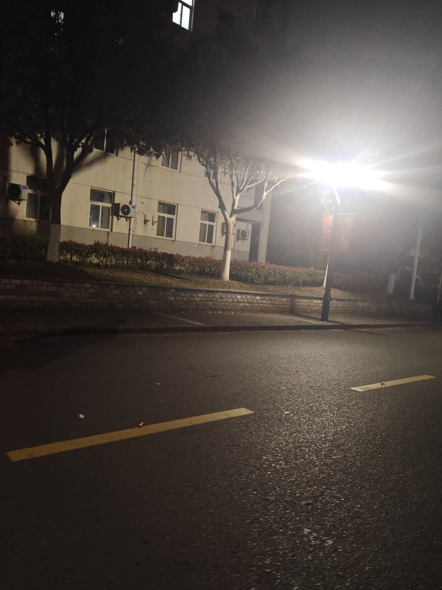 A nighttime shot of a street in Jiangning District, China. A two-story building with multiple windows and several air conditioning units attached to the exterior is situated behind a brick retaining wall and shrubbery. Two trees flank the building. A lamppost stands on the right side of the frame, emitting a bright light. The asphalt street in the foreground has yellow painted markings. The general environment is dark.