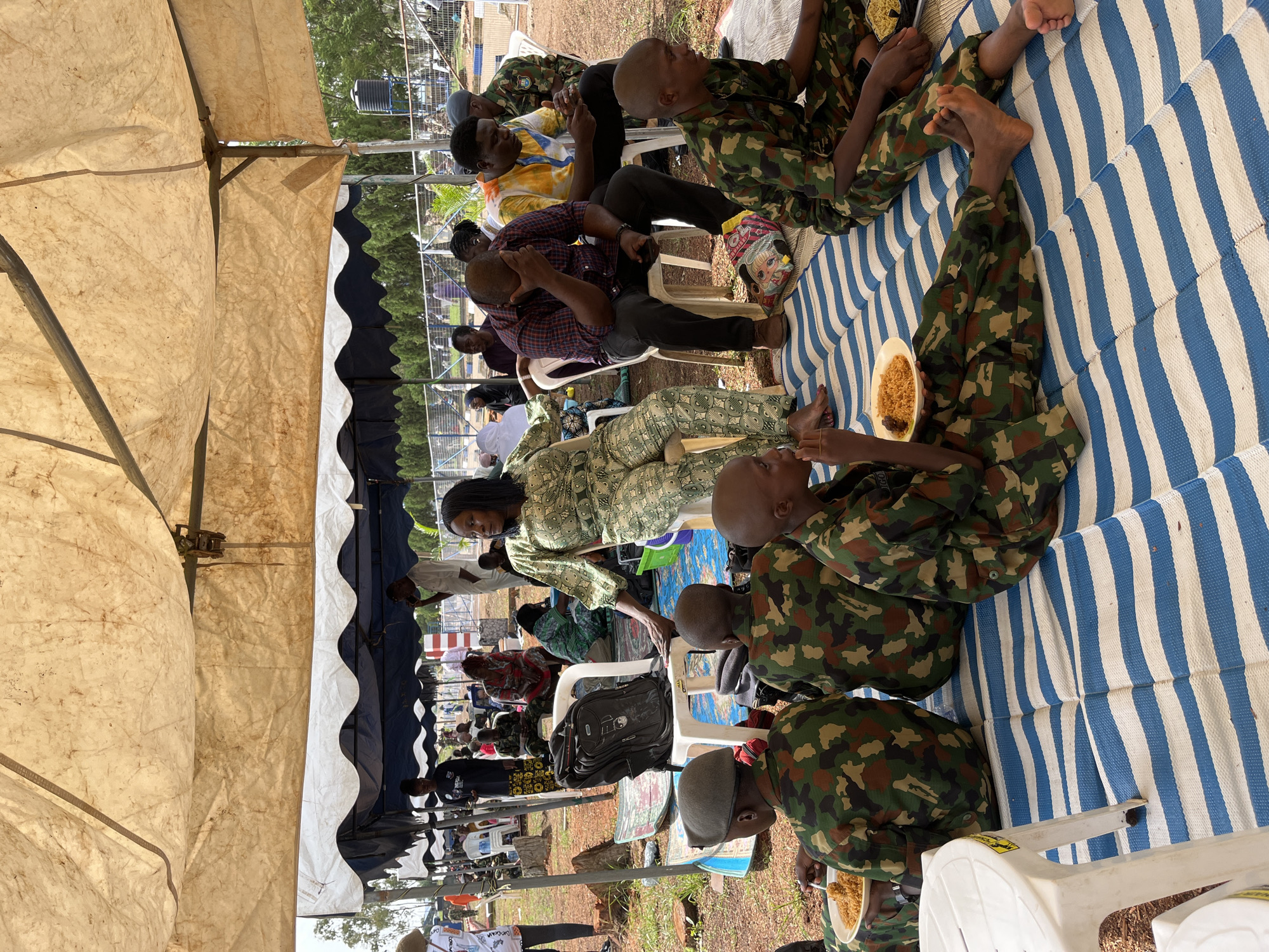 In Bar Jirgi Summa, Nigeria, a group of individuals are gathered under a large, makeshift tent. The tent's pale fabric roof is supported by metal poles, and its sides are open, revealing the surroundings. The ground is covered with a blue and white striped mat. Several people are seated on the mat, including at least five young individuals in camouflage military uniforms, each holding a plate of food. To the right, a woman in a green patterned dress is seated on a white plastic chair, looking towards the right. Behind her, several other people are seated on chairs and the ground, some talking and others appearing to be resting. In the background, trees and a fence are visible, suggesting an outdoor location within or near a settlement. A black backpack rests on a chair to the left of the woman in the green dress.