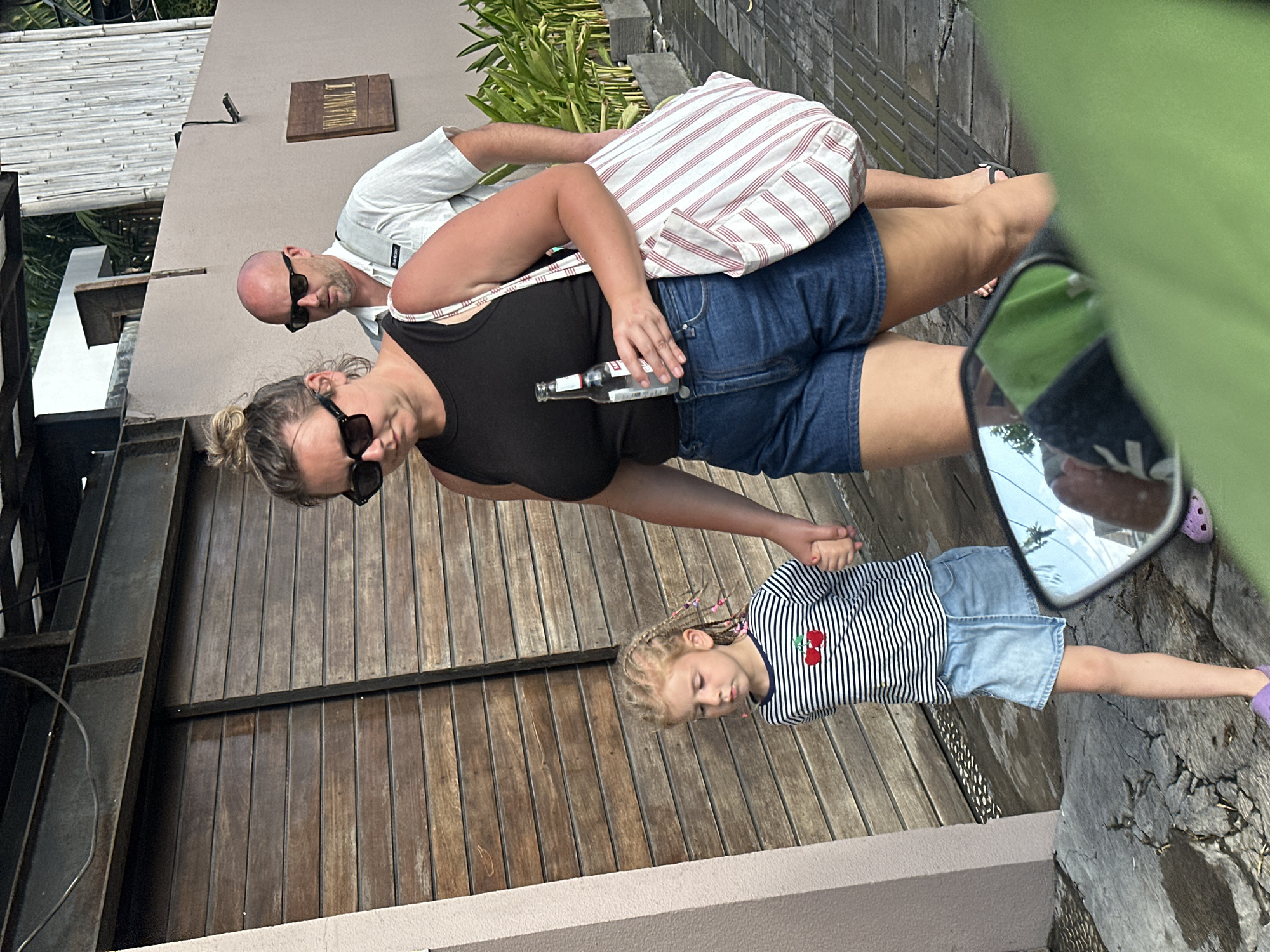The image shows three people in an outdoor setting. A man and a woman are standing, with the woman holding a bottle of what appears to be beer. A young girl with braided hair is also present, holding the woman's hand. The woman is wearing a black tank top and denim shorts, and the man is wearing a white short-sleeved shirt and sunglasses. The girl is dressed in a striped shirt and denim shorts.
Behind them is a building with wooden doors and a sign that reads "TULAMBEN" in gold lettering. The scene appears to be a pathway or courtyard, with wooden planks on the ground. The lighting suggests it is daytime, possibly late afternoon, with no obvious indications of weather conditions. The presence of a sign with a location name might suggest a resort or tourist area. The overall mood seems relaxed and casual. In the reflection of a mirror, a hand holding a phone is visible.