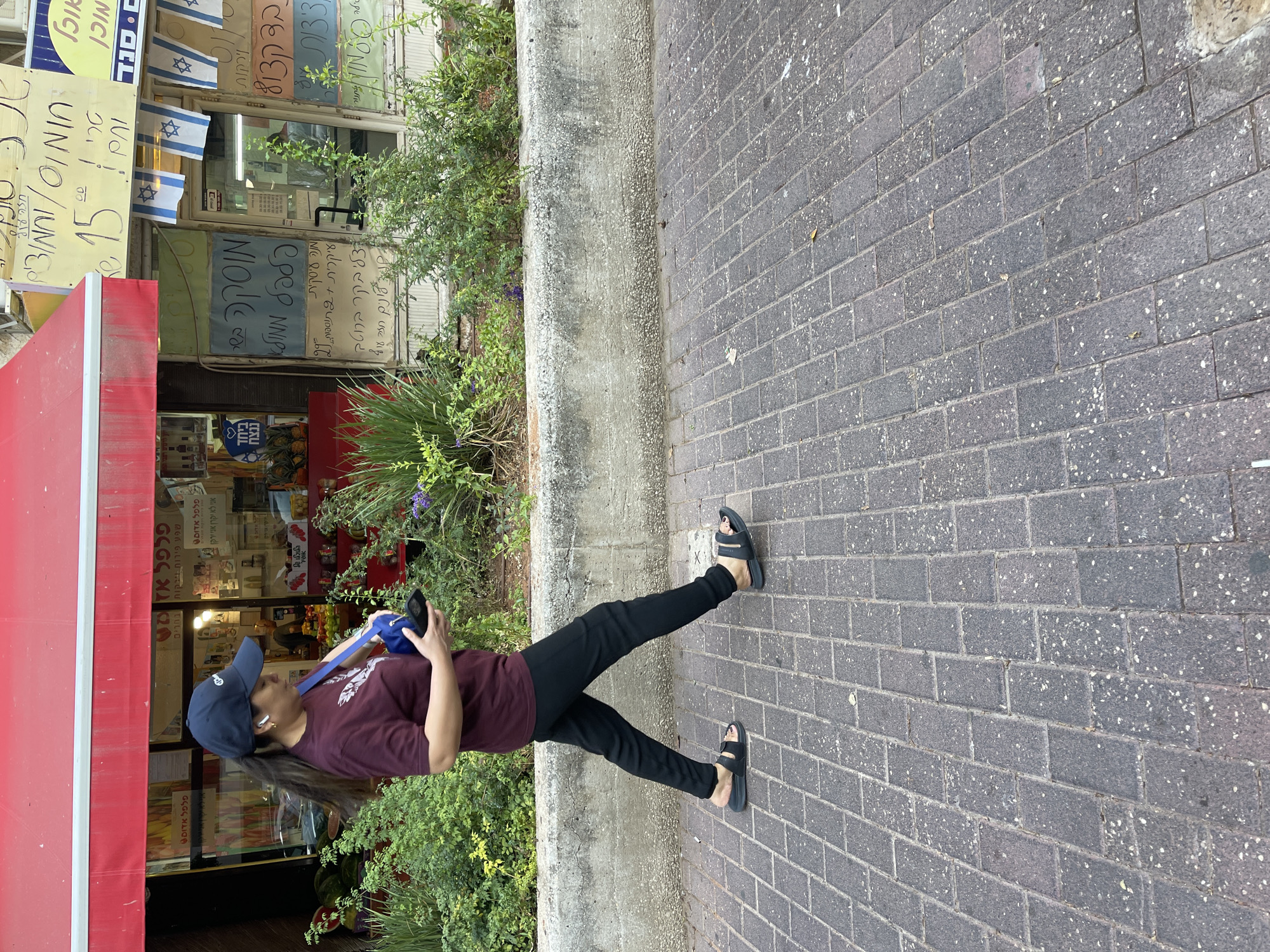 A full shot captures a woman walking on a cobblestone sidewalk in front of a shop in Kfar Saba, Israel. She is wearing a dark maroon t-shirt, black pants, black sandals, and a dark blue baseball cap. A blue purse is slung over her left shoulder, and she is holding a black mobile phone in her right hand. The shop's exterior features a red awning and windows displaying various goods, with signs written in Hebrew. Israeli flags are visible hanging from the storefront. Green plants and shrubs are planted along the sidewalk in front of the shop. The scene appears to be during daylight hours.