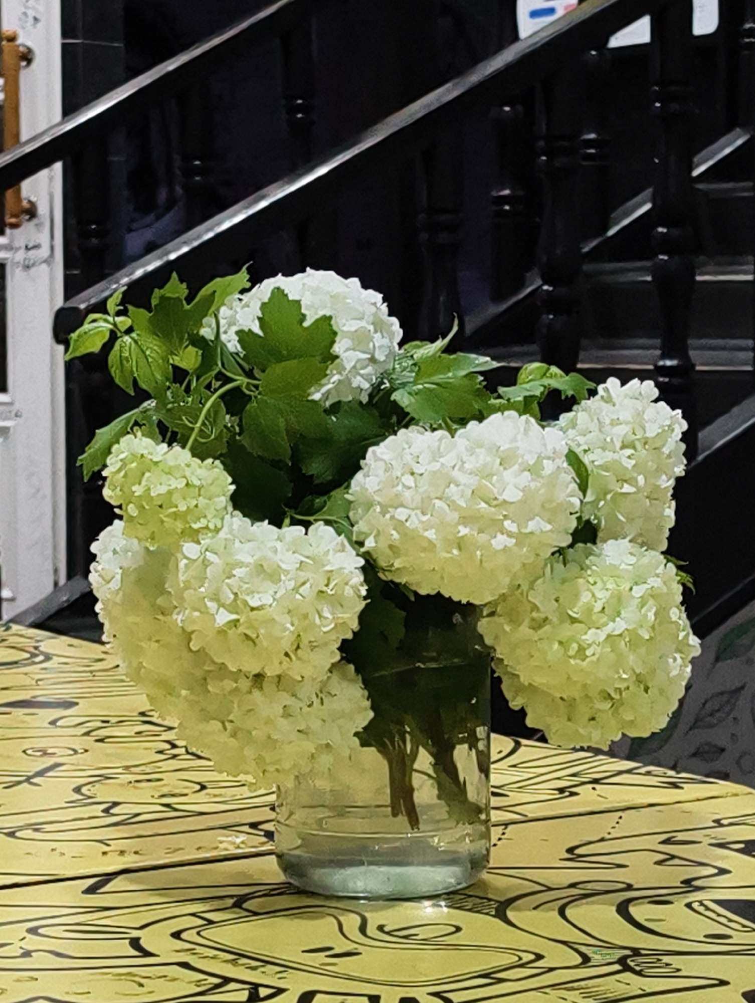 The image shows a bouquet of white hydrangea flowers in a clear glass vase on a table with a yellow and black patterned surface. The bouquet is lush with several flower heads and green leaves. Behind the flowers is a black wooden staircase with white trim. The photograph was taken in Tashkent, Uzbekistan.