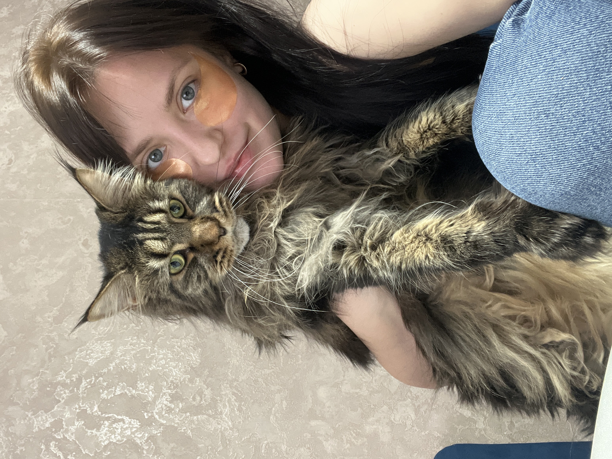 A woman with dark hair and fair skin is holding a large, brown tabby cat. She has what appear to be orange eye patches under her eyes. She is wearing blue jeans and gold hoop earrings. The background is a light-colored textured surface, possibly a countertop or floor. The location is Марковский, Russia.