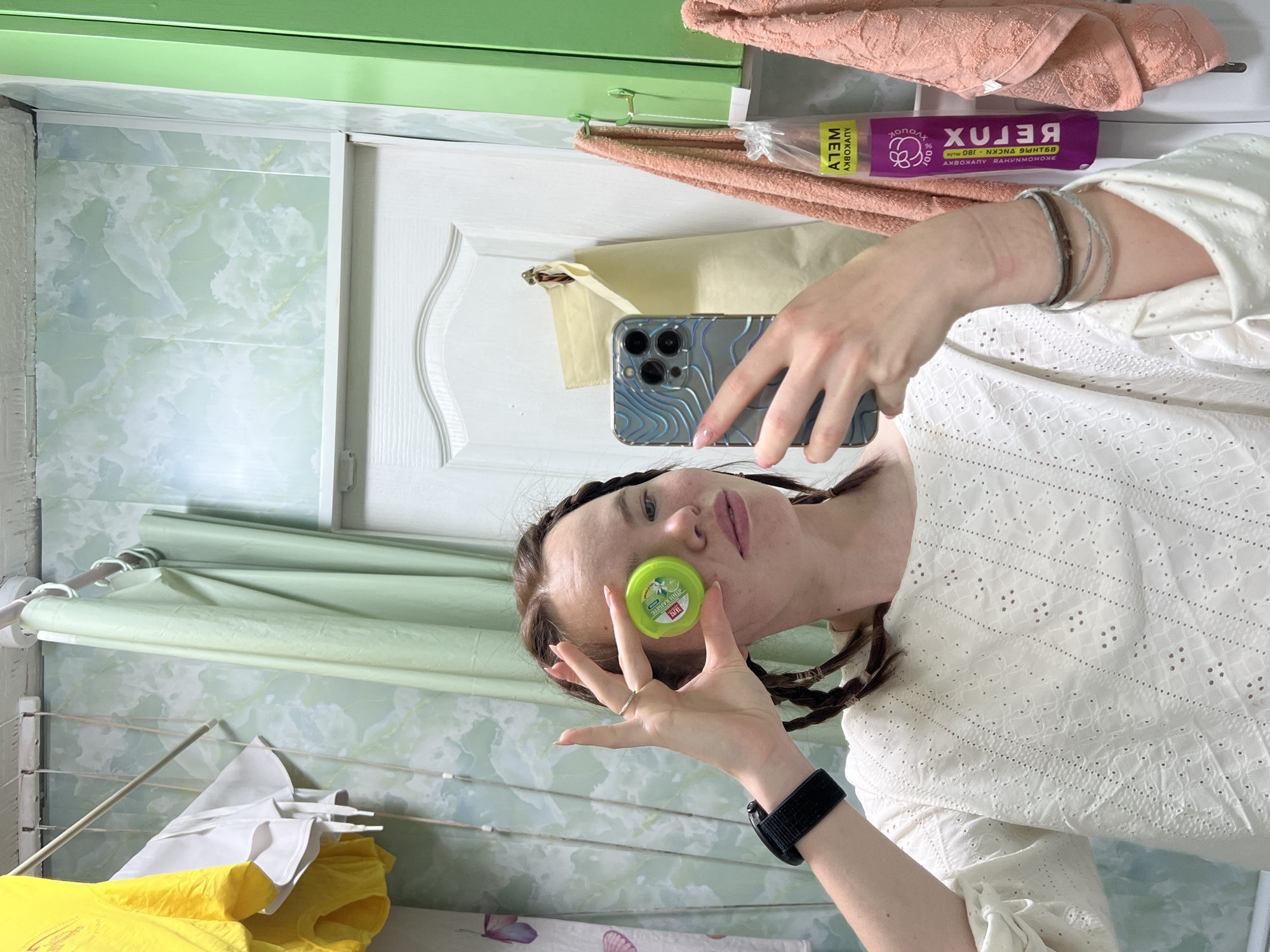 A person is taking a selfie with a smartphone in a bathroom. The person is holding a small, round, green container near their face. The background shows a bathroom interior with light green patterned walls, a white cabinet, and a clothes drying rack with clothes hanging on it. There is a towel rack above the cabinet, with a peach-colored towel hanging on it, along with a purple package. The person appears to be standing near a clothing rack with laundry on it. The image was taken in Chaikovsky, Russia.