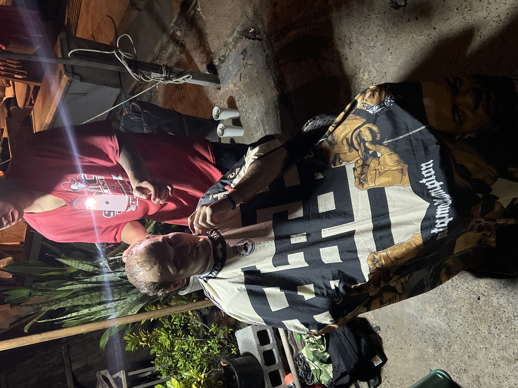 Scene: The photograph depicts two people under dim lighting conditions, possibly outdoors at night. The foreground features a person lying on a surface, covered with a black and white blanket that has graphic images and text on it. Another person, wearing a red shirt, stands above and holds a phone, possibly taking a photo.
People: One person is lying prone and covered with a distinctive blanket. The second individual, wearing a red shirt, is actively holding a mobile phone.
Objects: The scene contains a blanket, a mobile phone, plants, cement blocks, an empty trash can, and various other items scattered on the ground, including rope and what appear to be PVC pipes.
Actions: The person in the red shirt is using their phone, likely taking a photograph of the person lying under the blanket.
Interactions: There's an interaction between the two people as the person with the phone is presumably focused on the person lying down.
Environment: The location appears to be an outdoor area with concrete ground and plants. The lighting is poor, indicating a nighttime setting with limited artificial illumination.