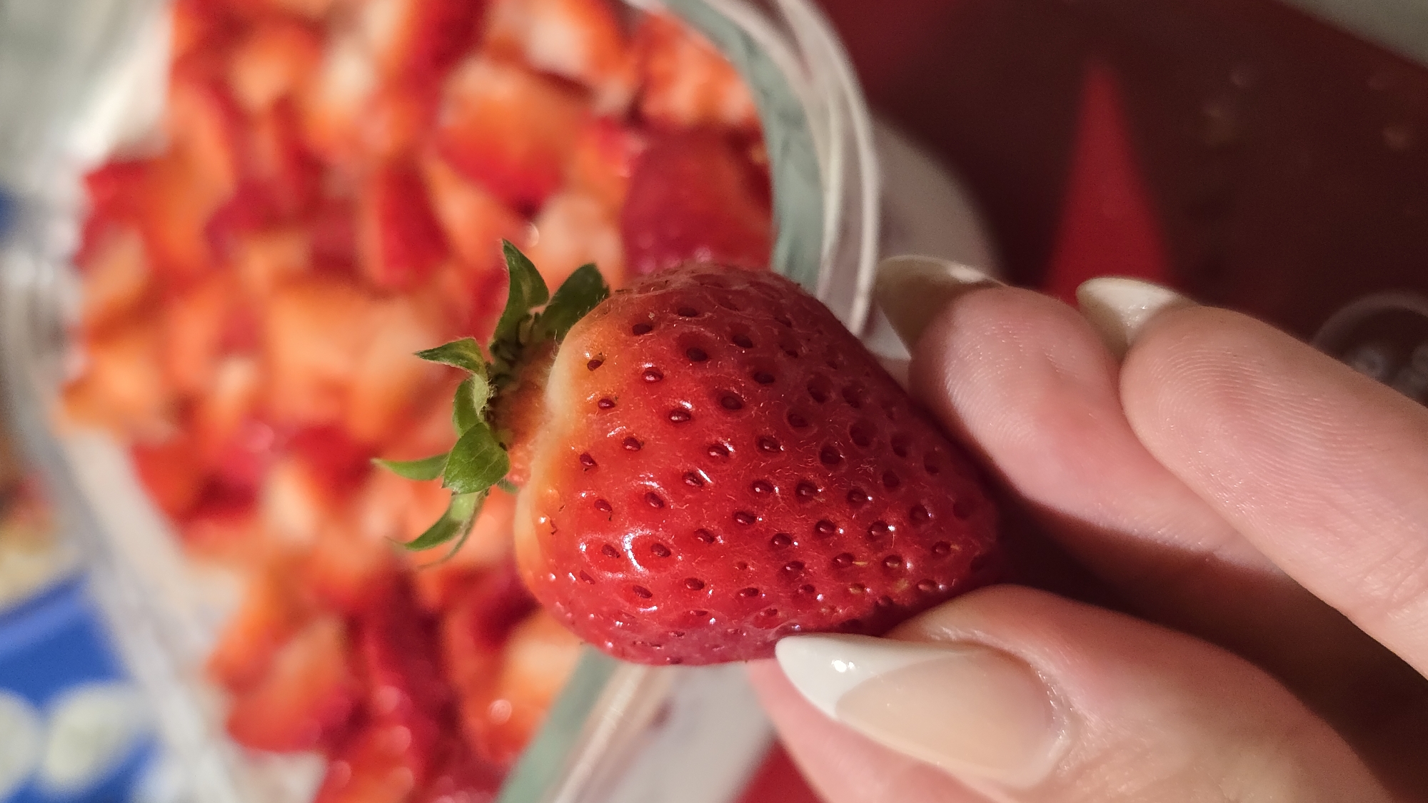A close-up shot shows a hand holding a strawberry in front of a container of chopped strawberries. The strawberry in the hand is red, with green leaves at the top. The fingers have manicured nails with a white tip design. The container behind is filled with pieces of strawberries. The location is unknown.