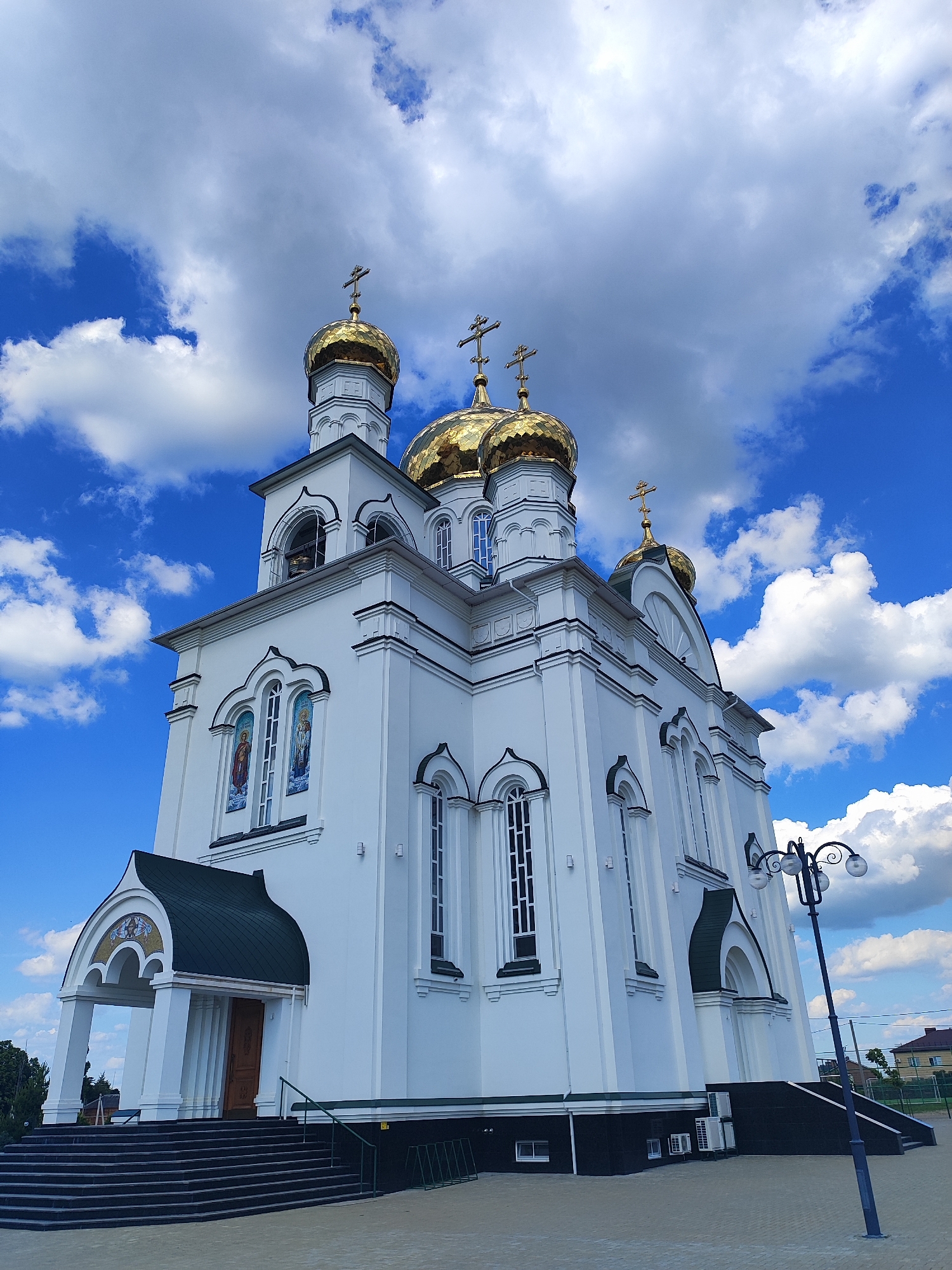 The image shows the Assumption of the Holy Virgin Cathedral in the city of Bryukhovetskaya, Russia. The cathedral is a white, multi-domed building with golden cupolas topped with crosses. The sky is blue with white, fluffy clouds. In the foreground, there is a paved courtyard with a lamppost and some steps leading up to the entrance of the cathedral. The architecture of the cathedral is ornate, with arched windows and decorative elements.