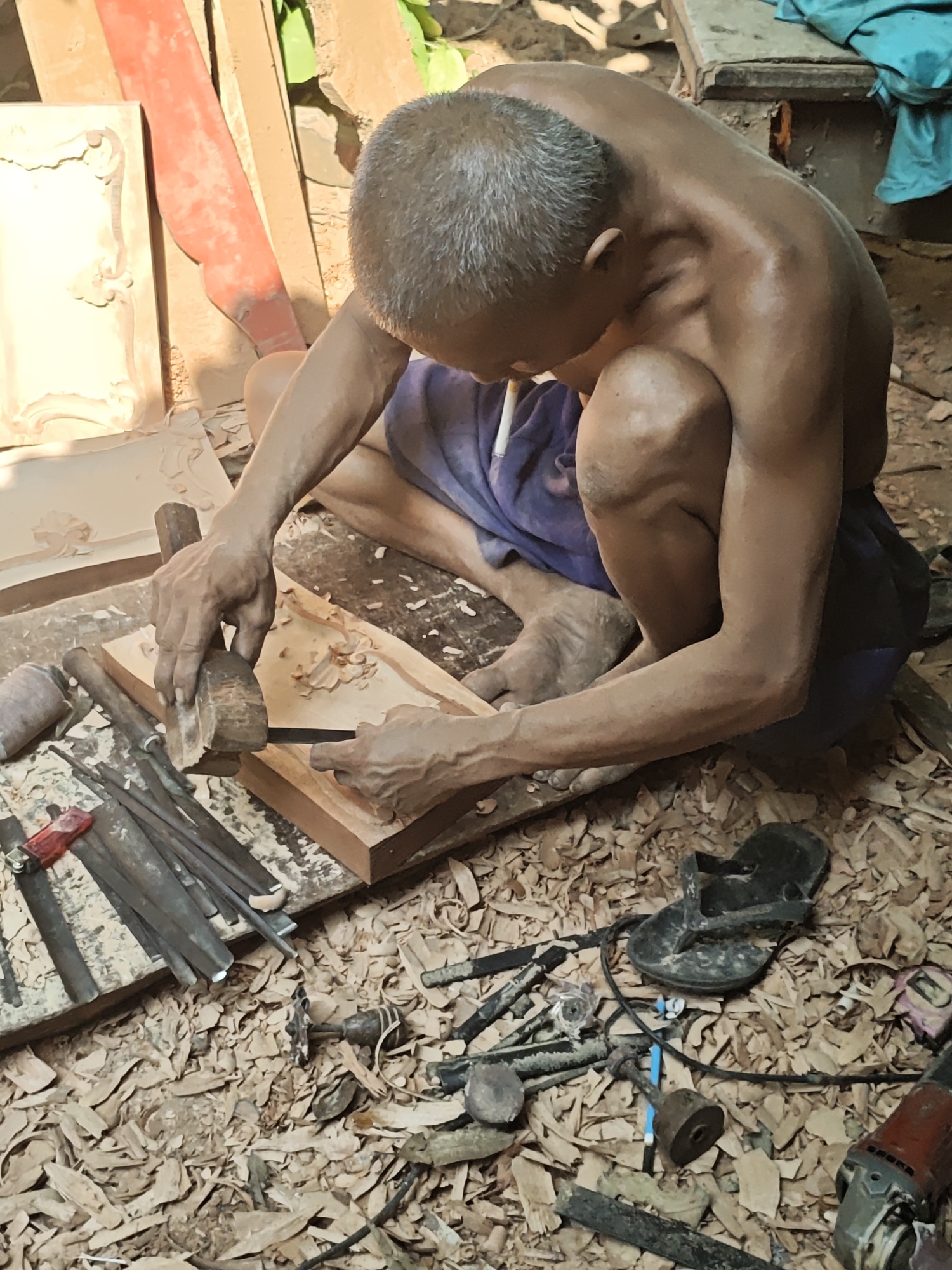 In Ngasem, Indonesia, an older man with graying hair sits on the ground, intensely focused on carving wood. He is shirtless, wearing blue shorts, and a cigarette dangles from his lips. His muscular arms and hands are visibly strained as he wields a chisel and hammer, shaping a piece of wood that rests on a larger plank. The ground around him is littered with wood shavings, testament to his work. Scattered among the shavings are various carving tools, including chisels and a small red lighter. A pair of black flip-flops lies nearby, and a power tool, possibly an angle grinder, is visible at the edge of the frame. The background shows more wooden materials, some unfinished carved pieces, and hints of a rustic, outdoor workshop environment.