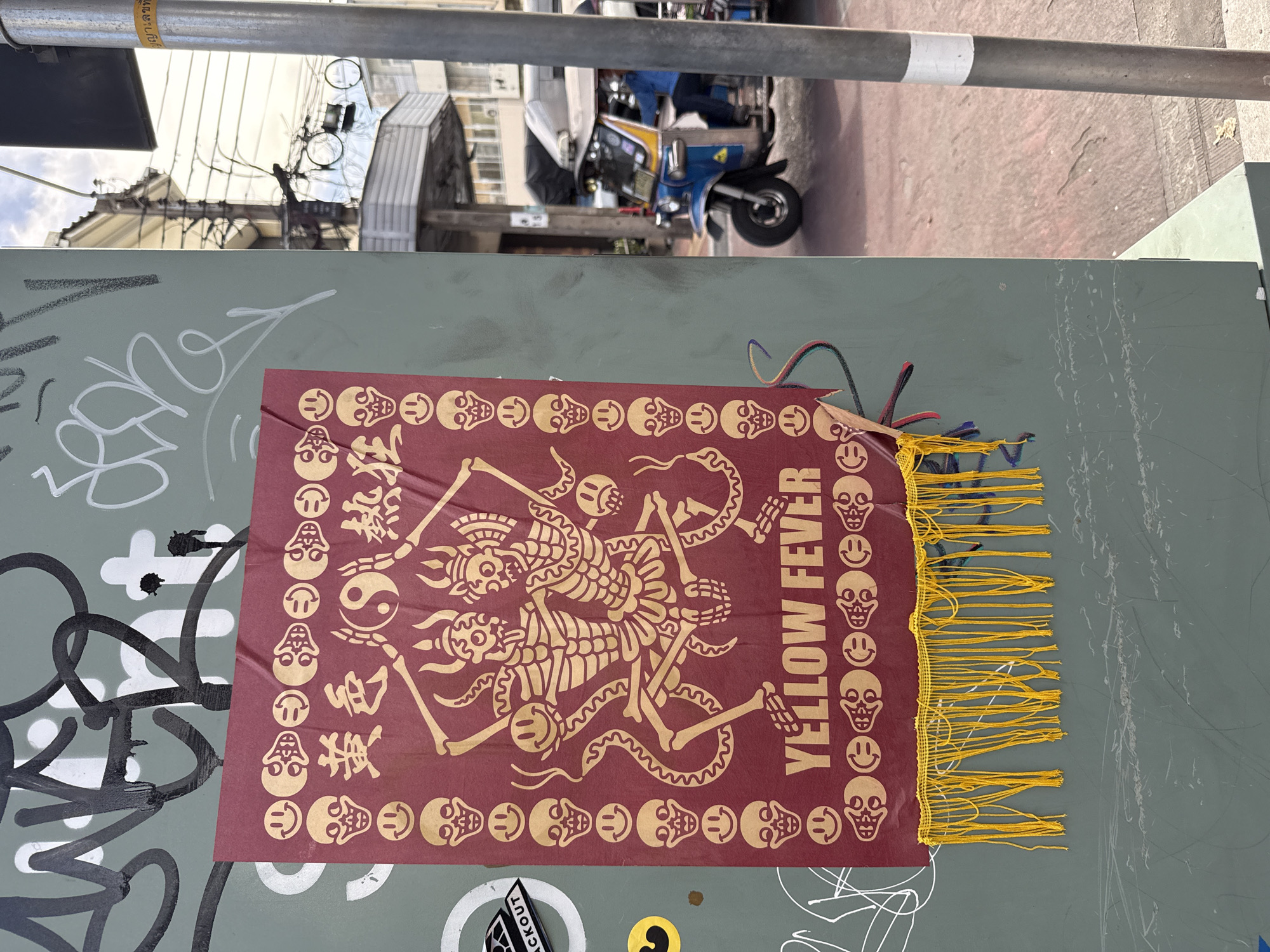 A close-up, medium shot shows a maroon banner with yellow designs attached to a gray, graffiti-covered metal surface. The banner, titled "YELLOW FEVER," features a graphic of intertwined skeletons and a decorative border of smiley face/skull images and snakes, along with Chinese characters and a Yin-Yang symbol. The banner has a fringe of yellow strings along one edge. The metal surface shows signs of wear and is marked with black and white graffiti. In the background, a partial view of a street in Bangkok, Thailand, includes an auto rickshaw (Tuk-Tuk) parked on the side of the road. The perspective is slightly tilted.