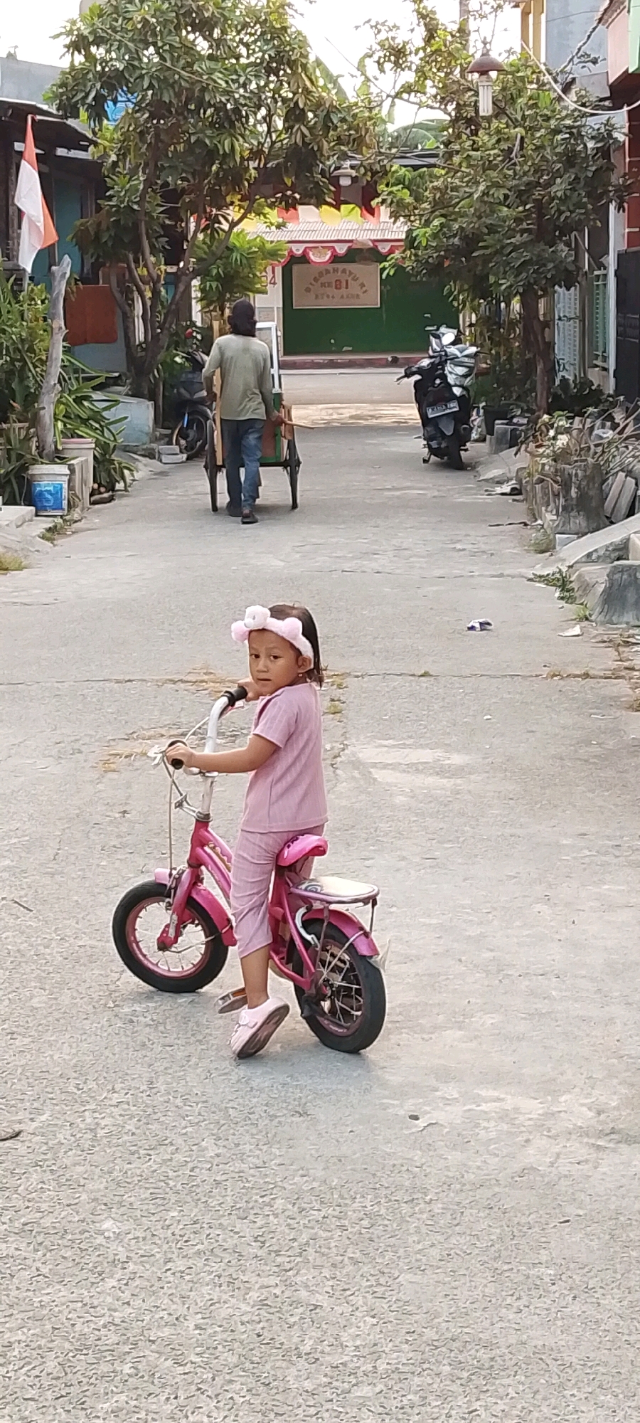 A young girl in a pink outfit and headband sits on a pink bicycle, looking at the camera. She is on a paved street, likely in Satriamekar, Indonesia, as indicated by the background elements such as houses and a motor scooter. The street appears to be in a residential area, with trees and some buildings visible. In the background, a man pushes a cart, and other motorbikes are parked. The overall scene depicts a bright, daytime outdoor environment.