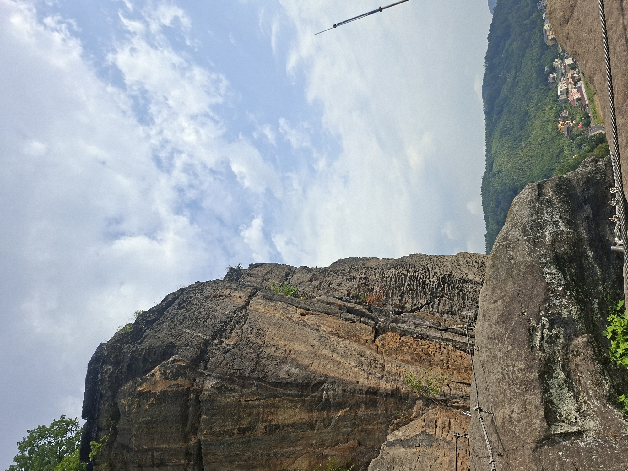 The image depicts a dramatic cliff face on the left, with a hiking path secured by cables and metal handrails traversing the rocky terrain. In the foreground, a large, weathered boulder is prominent, also featuring cable railings. Beyond the cliff and boulder, a verdant, forested hillside descends towards a village with scattered houses. A thin, dark pole extends vertically into the sky on the far right. The sky is partly cloudy with a bright, diffused light. The scene appears to be a scenic overlook or a section of a trail offering views of a valley and settlement. The location is identified as Děčín, Czechia, suggesting this is a view from a prominent geological feature within or near the city. There are no people visible in the image.