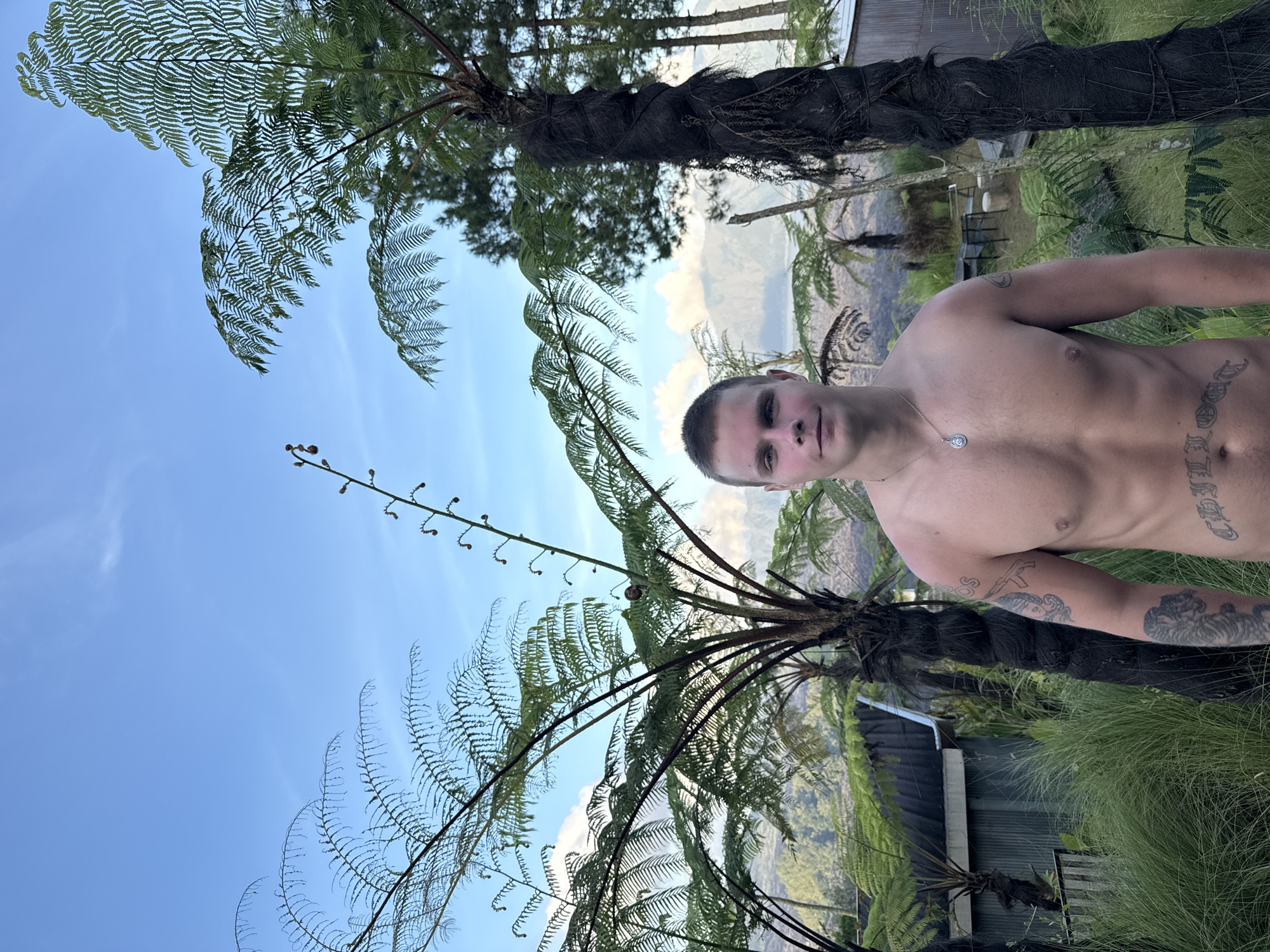 The image presents a shirtless male standing outdoors, seemingly in Kintamani, Indonesia, as suggested by the lush greenery and mountain backdrop. He is positioned amidst tree ferns, with a dark trunk and expansive fronds framing the scene. He is wearing a necklace with a circular pendant. His body features visible tattoos on his torso and arms. In the background, a building with a corrugated metal roof is visible. The environment indicates a mountainous region, likely a natural setting in Kintamani.