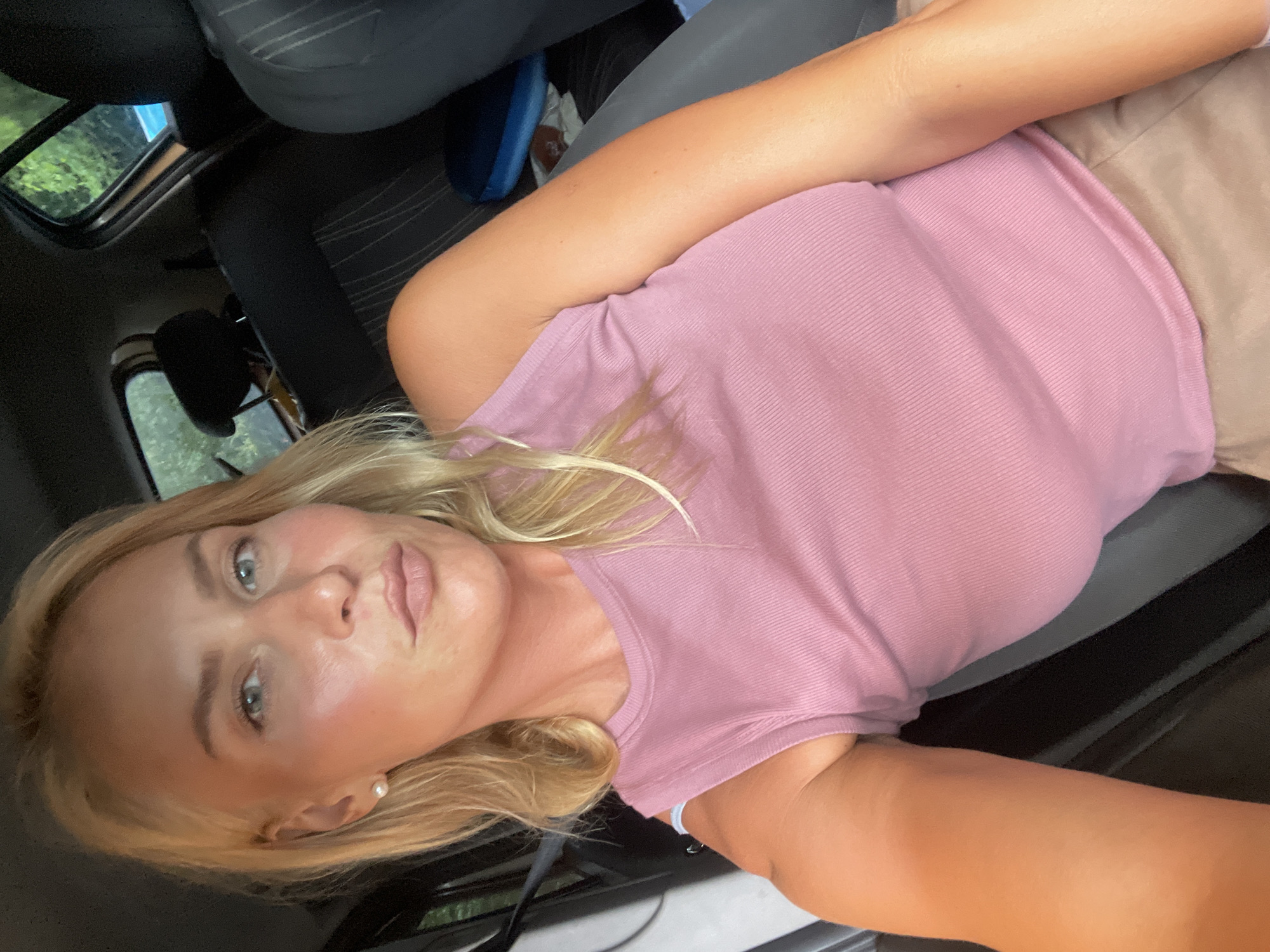 A blonde woman with light blue eyes and fair skin is seated in the passenger seat of a car, taking a selfie. She is wearing a pink ribbed tank top and light brown shorts. Her blonde hair is styled loosely around her shoulders. The interior of the car is visible, including the headrest of the driver's seat, part of the back seat, and the rear window, through which trees and foliage can be seen. The lighting suggests it is daytime, possibly overcast or in a shaded area. There are no other people visible in the image. The background context does not provide any specific indicators to pinpoint the location within Baranovka, Russia.