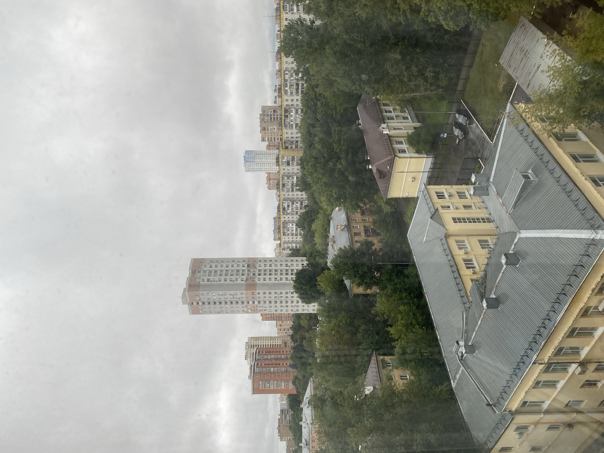 An overcast day in Odintsovo, Russia, is depicted from a high vantage point looking out over a dense urban landscape. The foreground features the grey, corrugated metal roofs of several buildings, one a light yellow structure with multiple windows and air conditioning units visible. Adjacent to it, another building also exhibits a yellow facade. Beyond these, a dense canopy of green trees dominates the midground.
In the background, a mix of apartment buildings and taller, modern structures punctuate the skyline. Prominent among them is a very tall, light-colored apartment building with decorative reddish-brown accents near its peak. To its left, a shorter, reddish-brown brick apartment building stands out. Further in the distance, a series of multi-story residential buildings with repetitive window patterns stretch across the horizon.
The sky is a uniform, pale grey, suggesting cloudy weather, possibly a light rain or drizzle, as evidenced by the damp appearance of the asphalt in the midground parking area where two dark-colored cars are parked. The overall impression is one of a typical residential area under a muted sky.