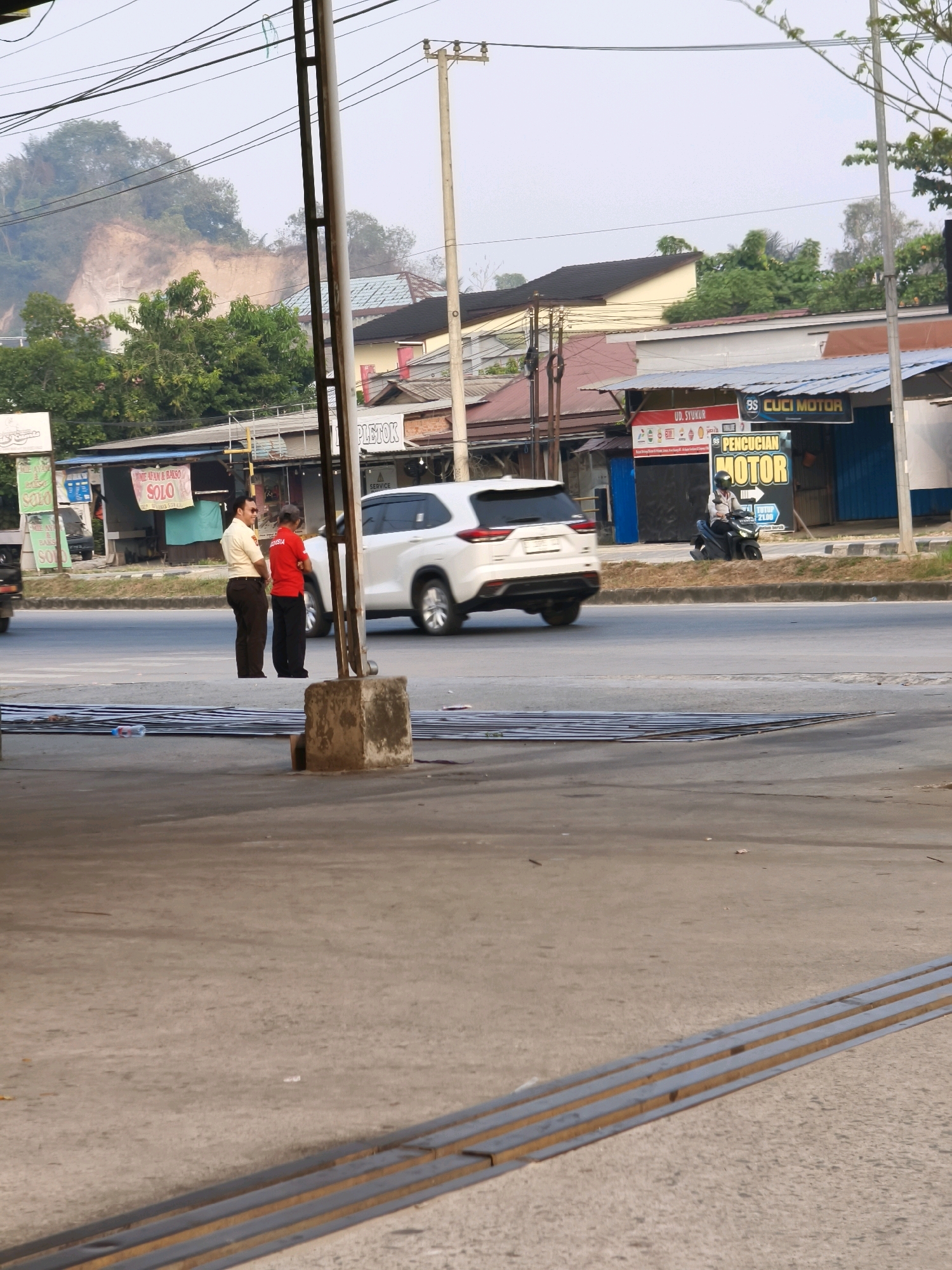 This is a medium shot of a street scene in Samarinda, Indonesia. Two men are standing on the side of the road, seemingly waiting. A white SUV is driving on the road behind them. In the background, there are several buildings, some of which appear to be shops. A motorcycle is parked in front of one of the shops, and a person is sitting on it. The overall environment appears to be urban, with overhead power lines and trees visible. The road has a median with grass. The foreground shows a concrete surface with some metal bars laid out. The time of day appears to be late afternoon or early evening, given the lighting conditions.