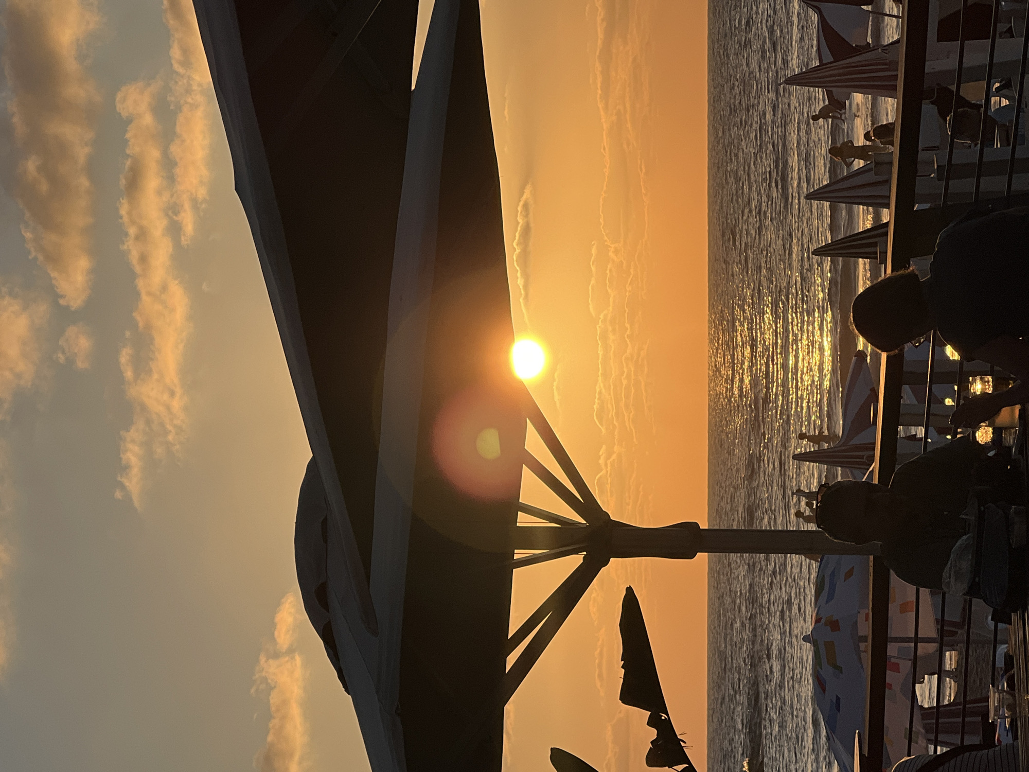 The image captures a serene sunset scene at a beach in Tel Aviv, Israel. Silhouetted against the vibrant orange and yellow hues of the sky, a large umbrella structure dominates the foreground, its dark form casting a stark contrast. Beyond it, the sun, a bright orb, sits low on the horizon, casting a shimmering golden path across the rippling surface of the sea.
Several beach umbrellas, some with striped patterns and others with colorful geometric designs, are visible further down the beach, suggesting a lively atmosphere even as the day winds down. In the mid-ground, a railing separates a seating area from the beach, where several people are enjoying the view. Their figures are largely obscured by shadow, but the overall impression is one of relaxation and enjoyment.
The clouds in the sky are wispy and tinged with the warm colors of the sunset, indicating fair weather and the approaching evening. The overall mood is peaceful and picturesque, evoking a sense of calm and beauty associated with a beach sunset. There is no visible text in the image.