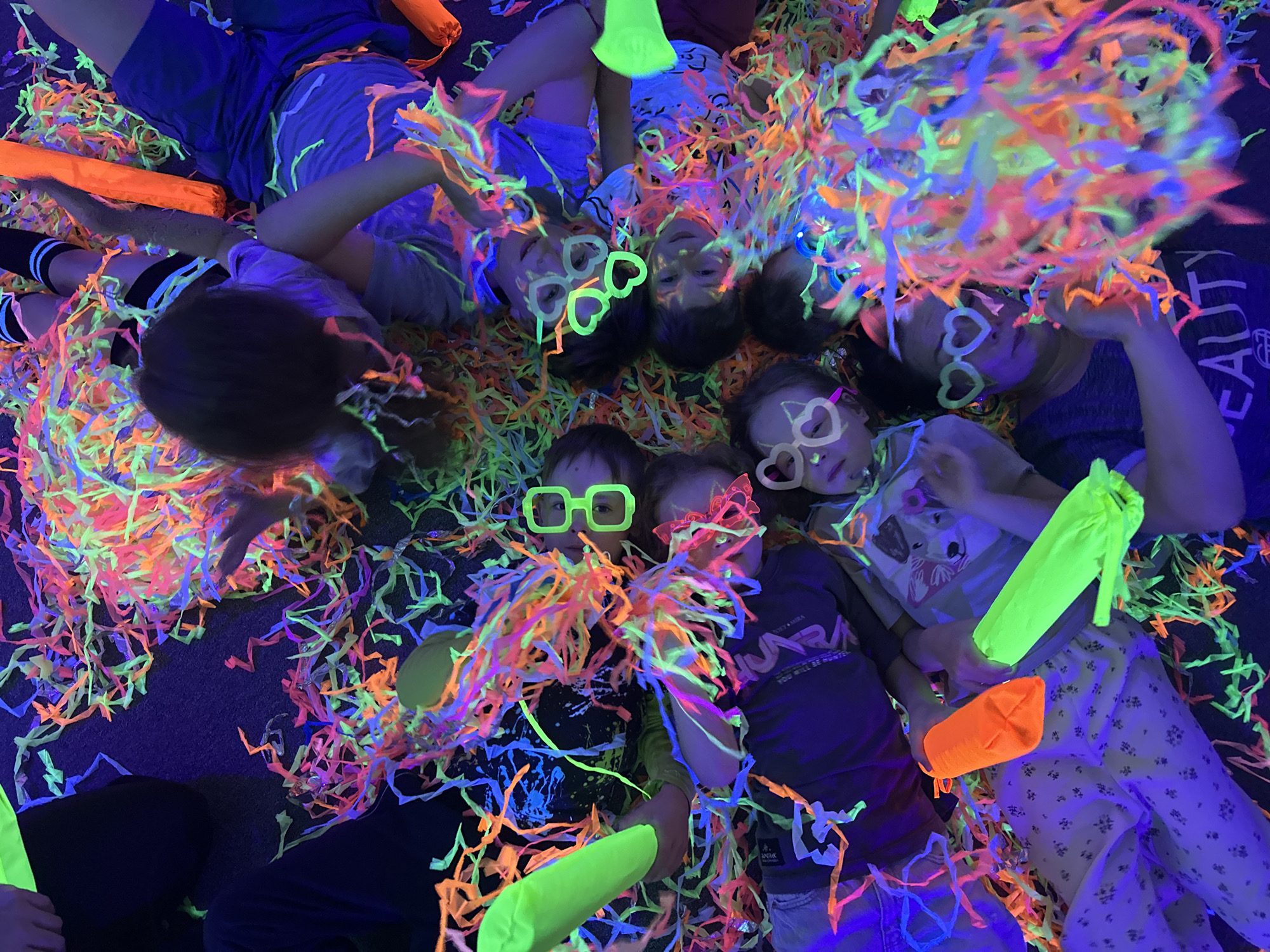 The image captures a group of eight individuals lying on what appears to be a dark-colored carpet in a room illuminated by black lights. Confetti lit with black light is scattered all around the children and adults. Everyone is wearing glow-in-the-dark glasses with different shapes such as hearts and rectangles. There are also several glowing neon green and orange objects or toys among them. The age range appears to be from small children to adults. Some are wearing dark clothing while others have on light-colored or printed shirts and pants. The location is unknown.