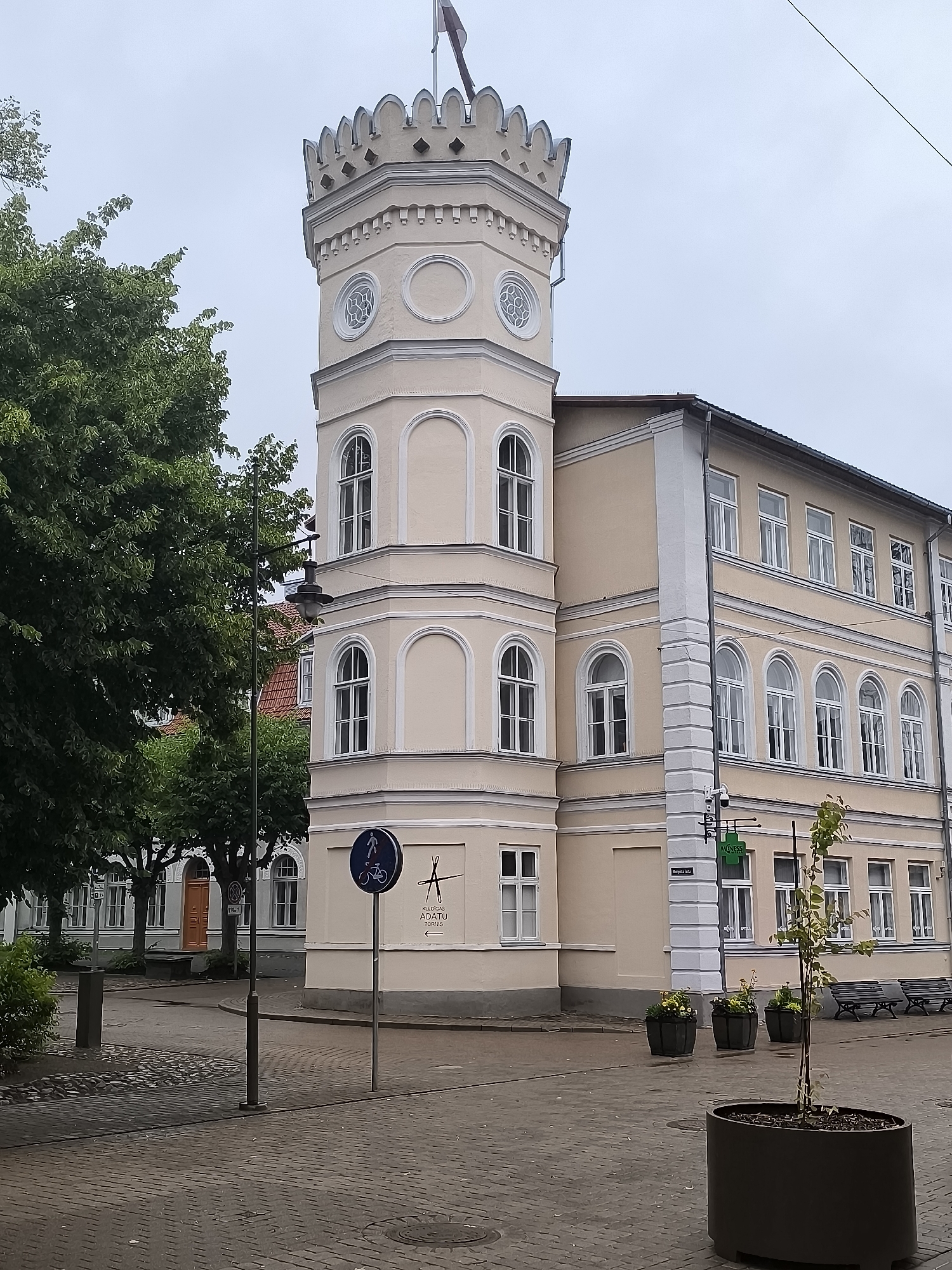 The image depicts the prominent, pale yellow building in Kuldīga, Latvia, located at a street intersection. The building features a distinctive crenellated tower with decorative circular windows and arched windows on its lower floors. The main facade of the building is a pale yellow with white trim around the windows and cornices. Several arched windows with white frames are visible on the upper floors of the main building. A street sign with a pedestrian and bicycle icon is positioned on the sidewalk in front of the building. Further down the street, a pharmacy sign with a green cross is visible. Paved sidewalks and cobblestone streets are present. Trees line the street, and benches are visible in the background. The sky is overcast. There are no people visible in the immediate scene.