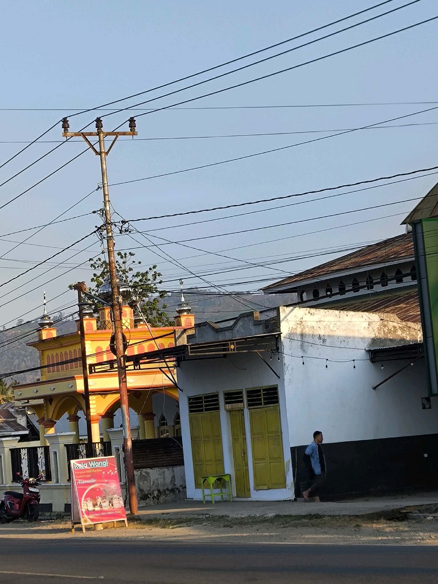 The image depicts a street scene in Tenilo, Indonesia, under a clear sky. A prominent feature is an old, weathered utility pole with multiple wires running across it, dominating the upper left quadrant. To the left of the pole is the facade of a brightly colored orange and yellow building, possibly a mosque, with arched entrances and decorative elements. In front of this building, a red scooter is parked. A white advertising stand for "Raja Wangi" is also visible.
To the right of the utility pole, a white building with yellow double doors and barred windows stands. A man in a blue shirt and dark pants is walking along the sidewalk in front of this building. The building's wall is partially illuminated by sunlight. Behind these buildings, a structure with a rusty, corrugated metal roof and a series of small arched openings is visible. The scene suggests a commercial or residential area.