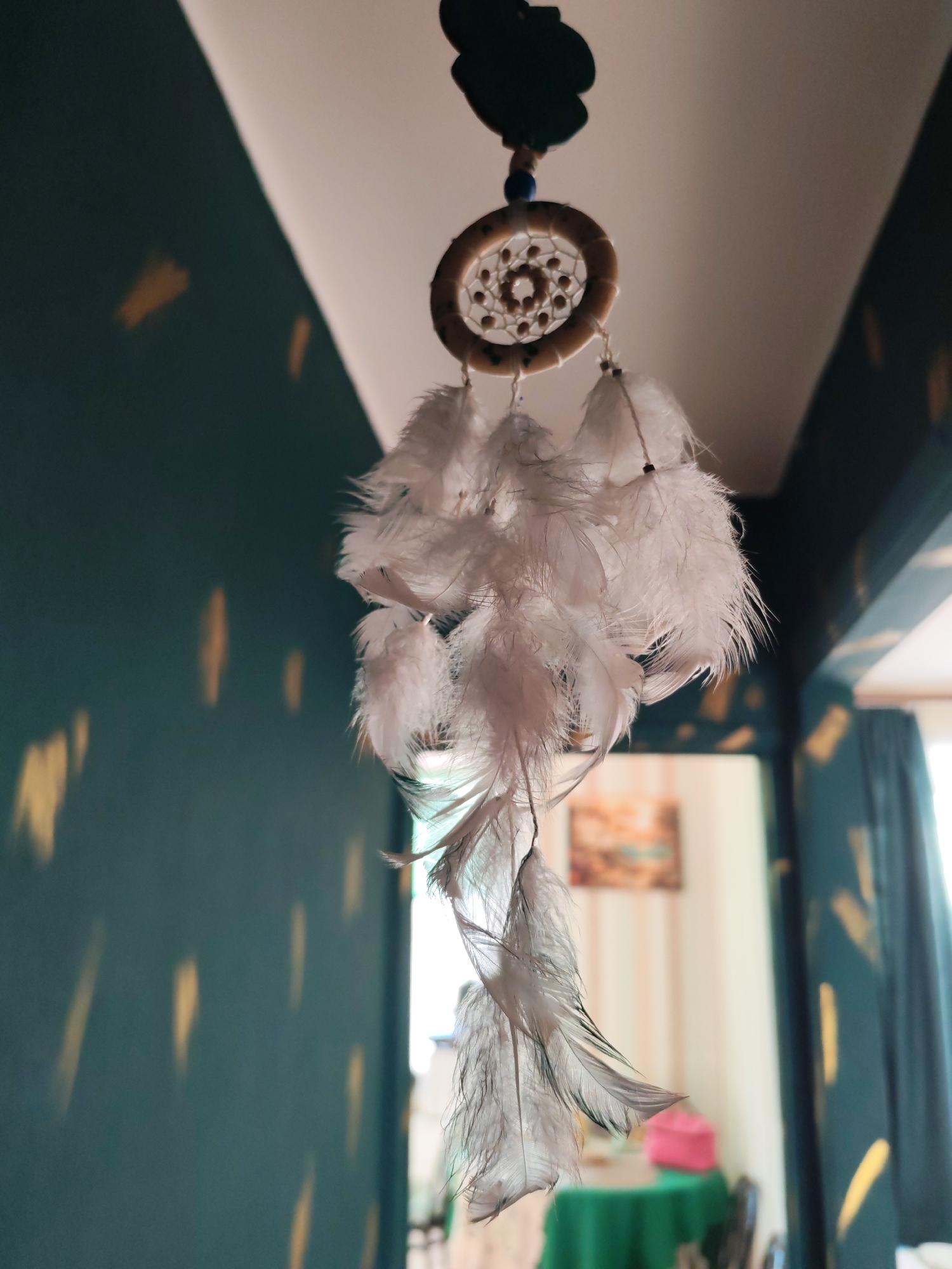 A dreamcatcher hangs in an interior setting. The wall is dark green with gold brushstroke-like marks. There is a doorway leading to another room with striped wallpaper and a green table. Light comes in through a window with dark green curtains. There is no evidence to indicate the location of the photograph as Izhevsk, Russia.