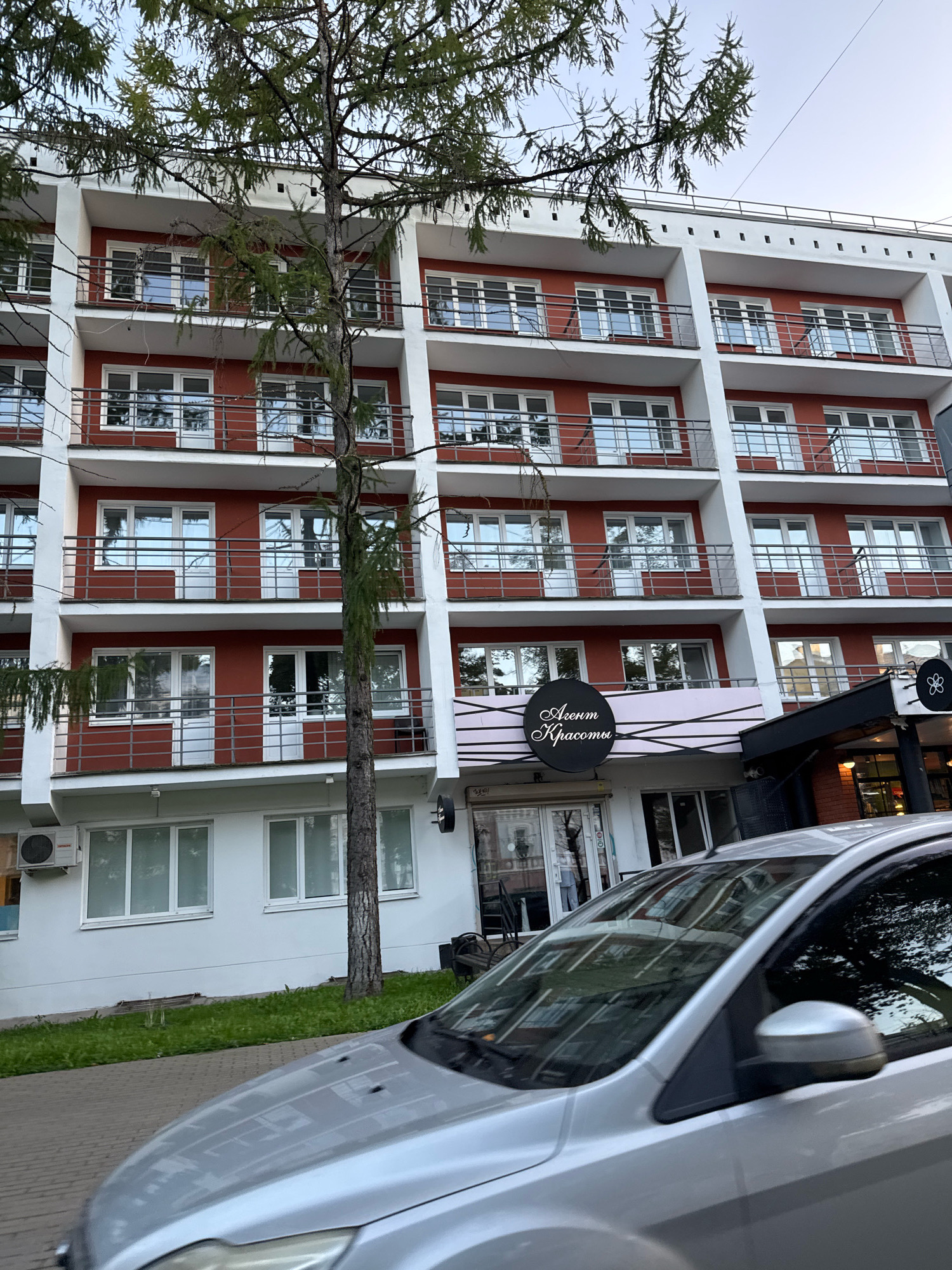 The image shows a modern building with a red facade and white balconies, likely an apartment building or a hotel. A tall, slender tree stands in front of the building, partially obscuring some of the windows and balconies. In the foreground, the hood and windshield of a silver car are visible, reflecting the building and the tree. The car appears to be in motion, suggesting the photo was taken from another vehicle. The building has a ground-floor entrance area with signage that reads "Агентство Красоты" (Agency of Beauty) in stylized script. The environment suggests an urban setting, possibly a street in the city of Tver, Russia, given the presence of a typical multi-story residential or commercial building and a car. The lighting indicates it's daytime, possibly late afternoon or early evening given the soft light and the reflections.