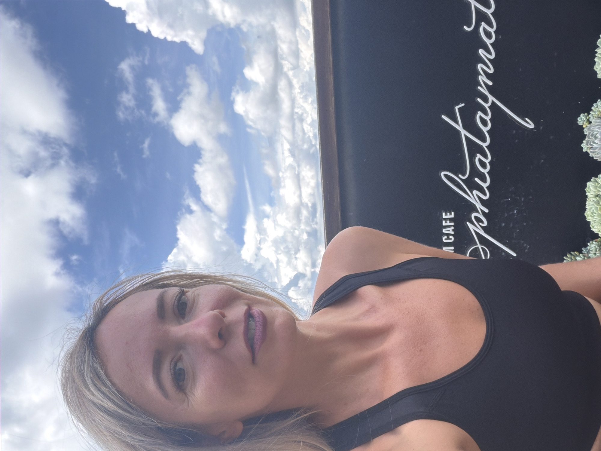 A woman with blonde hair and purple lipstick poses for a selfie outdoors. She is wearing a black sports bra. Behind her, a black sign with white cursive lettering reads "Sophiataymat Cafe". The sky is blue with scattered white clouds, and some succulents are visible at the bottom of the frame. The background suggests the image was taken in Da Lat, Vietnam.
