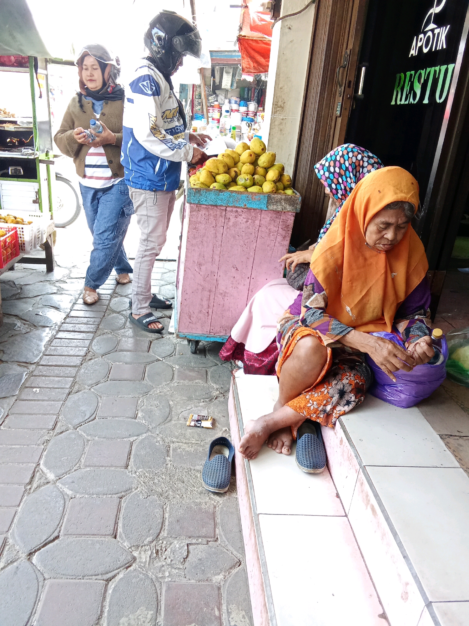 The image depicts an outdoor scene, likely a street market or roadside vendor area, in Banjarnegara, Indonesia. In the foreground, an elderly woman wearing an orange hijab and a patterned dress is seated on a low concrete ledge, looking down at something in her hands. Beside her, a large purple plastic bag rests on the ledge. To the left and behind her, another woman in a headscarf and a patterned shawl is partially visible. Further back, two individuals are standing near a cart piled with mangoes. One person, wearing a blue and white jacket and a helmet, appears to be handling some items on the cart. Another person, wearing a brown cardigan and a helmet, holds a water bottle. The ground is paved with textured rectangular stones. The background shows a building with a wooden door and signage that reads "APOTIK" and "RESTU". The overall impression is one of everyday commerce in an Indonesian urban setting.