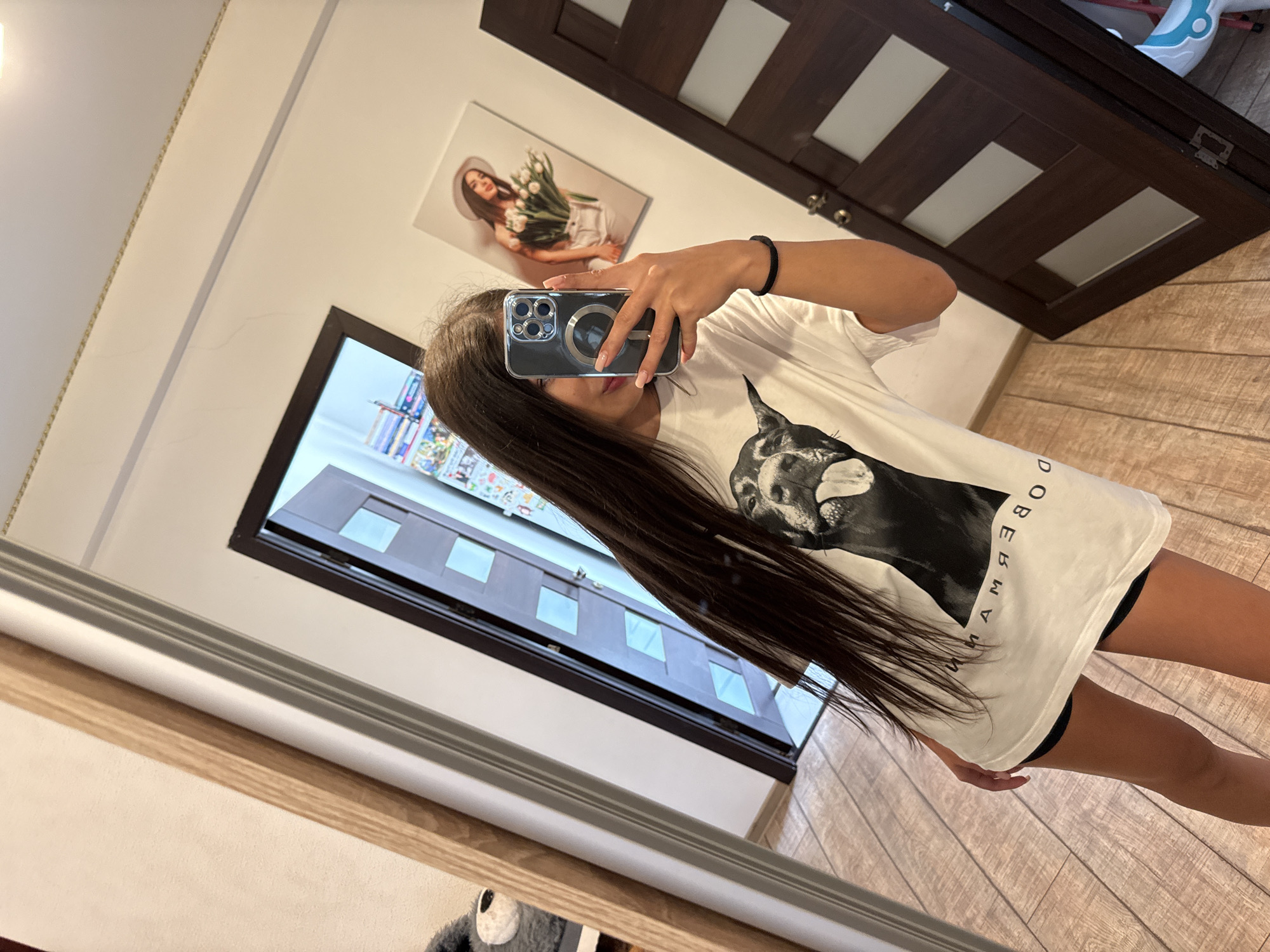 A young woman with long dark hair is taking a selfie in a room. She is wearing a white t-shirt with a picture of a Doberman dog on it and black shorts. The room has wooden floors and light-colored walls. There is a mirror to her left, reflecting her and the room. In the background, there are two doors, one slightly ajar, revealing a glimpse of books and posters on a shelf. A framed picture of a woman holding flowers hangs on the wall. The lighting in the room appears to be artificial. The image does not provide any specific information to determine its location in Chișinău, Moldova.