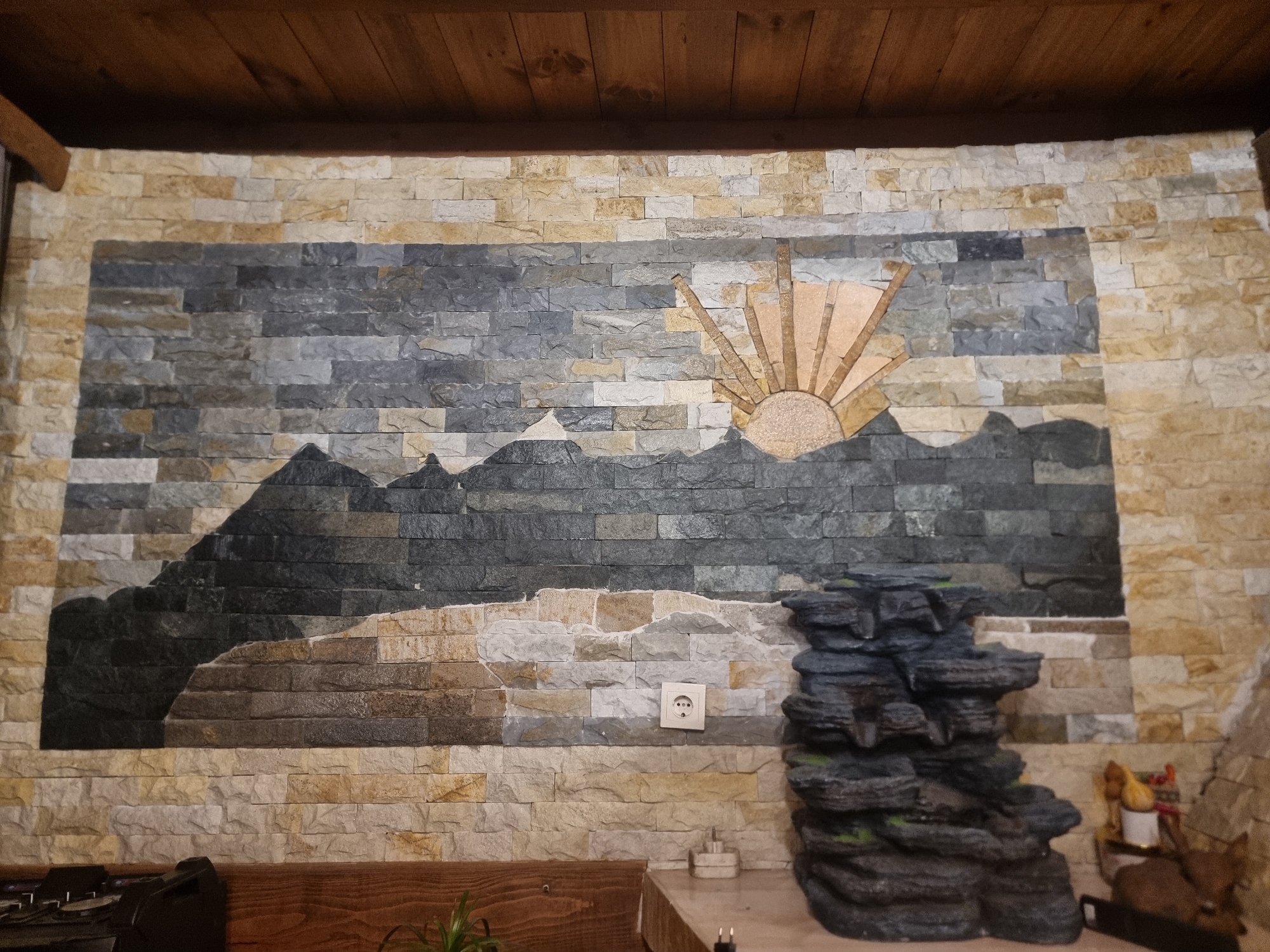 The image depicts an interior space, likely a home or business, in Banichan, Bulgaria. A large mosaic mural made of various shades of stone tiles dominates the central wall. The mosaic portrays a landscape featuring mountains, a sunrise with rays, and a body of water. The surrounding wall is also covered with horizontally stacked, light-colored stone tiles. Below the mural on the right side is a multi-tiered artificial rock formation, possibly a water fountain, with some green moss-like vegetation. An electrical outlet is visible on the wall to the left of the rock formation. In the bottom foreground, portions of wooden furniture are visible. The ceiling is made of dark wooden planks. To the right of the rock formation, on a lower level, are a couple of small figurines.