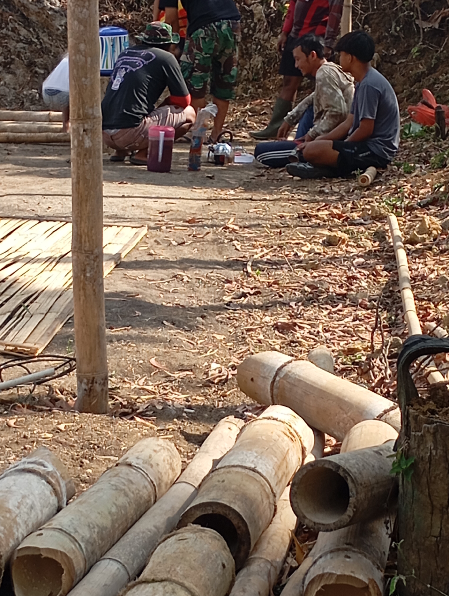In Ketepung, Indonesia, a group of individuals are gathered outdoors. In the foreground, several large sections of bamboo are laid out on the ground. To the left, a bamboo pole stands vertically. In the mid-ground, a person wearing a black t-shirt and a camouflage hat is crouching down near a large purple container. Other individuals are seated on the ground to the right, one wearing a camouflage long-sleeved shirt and another in a grey t-shirt. The environment appears to be dry and dusty, with fallen leaves scattered across the ground. A partially constructed bamboo structure or platform is visible in the background.