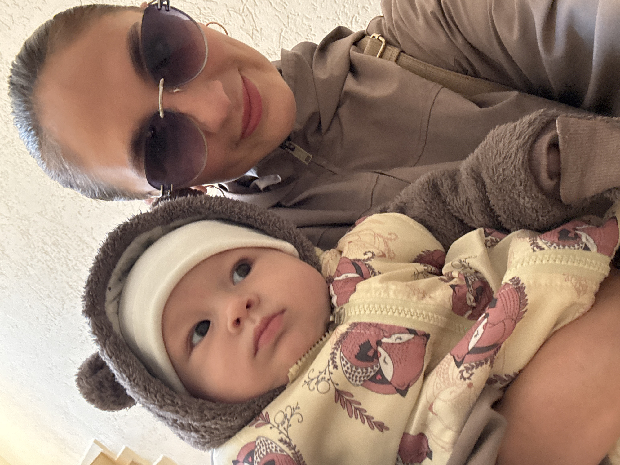 This low-angle selfie captures an adult woman holding an infant in a casual indoor setting. The woman occupies the upper portion of the frame, smiling warmly at the camera. She wears large, dark sunglasses and a neutral-toned (greyish-brown or taupe) hooded jacket, with a beige shoulder strap visible. Her lips are a soft pink.
Below and slightly to the left of the woman, the infant is held in her arms. The baby looks upwards and to the right, away from the camera, with a neutral and curious expression. The infant is dressed in a fluffy, dark brown hooded outfit with small bear-like ears, worn over a plain white hat. A cream or yellowish overall or jacket, patterned with reddish-brown stylized fox-like creatures and botanical motifs, is visible beneath the fluffy hood.
The background consists of a bright, textured surface, likely an indoor wall or ceiling. The lighting is diffuse and bright, suggesting a daytime scene. The image portrays a tender moment between the woman and child, conveying happiness from the adult's smile. No visible text is present in the image. The location context provided is Sochi, Russia.