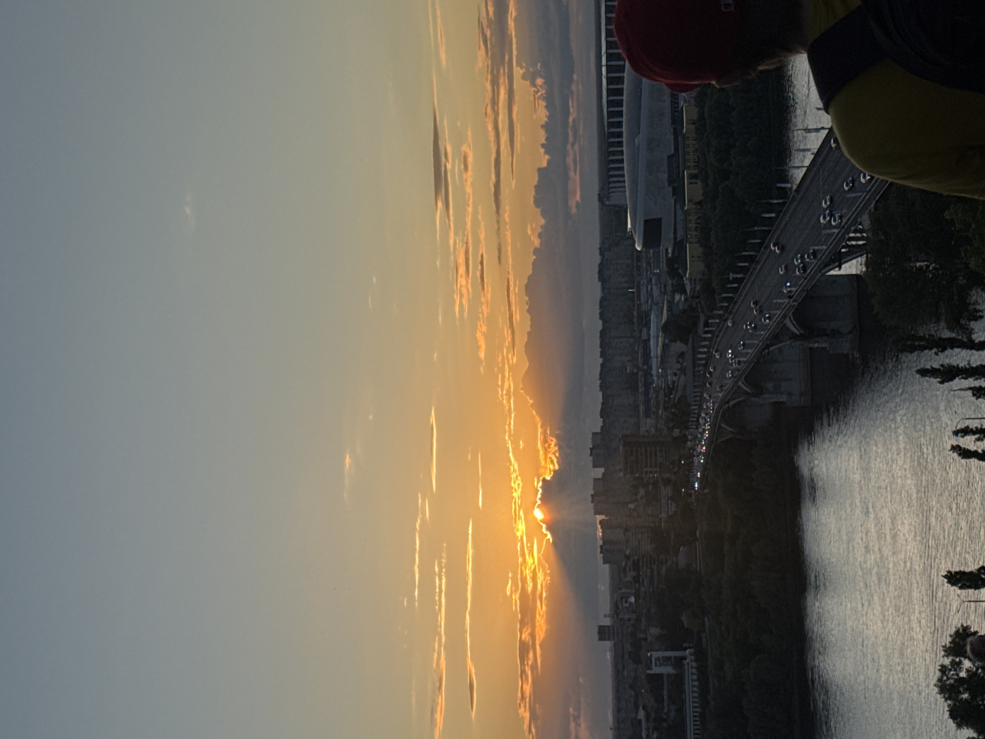 The image shows a sunset over the city of Nizhny Novgorod, Russia, as viewed from above. The sun is setting behind clouds, casting an orange glow across the sky and illuminating the clouds with golden light. A bridge spans a wide river in the foreground, with cars visible on the bridge. The city skyline is visible in the background, with numerous buildings and a stadium. A person wearing a red hat and yellow shirt is facing away from the camera, looking out at the view. The overall scene depicts a serene evening in the city.