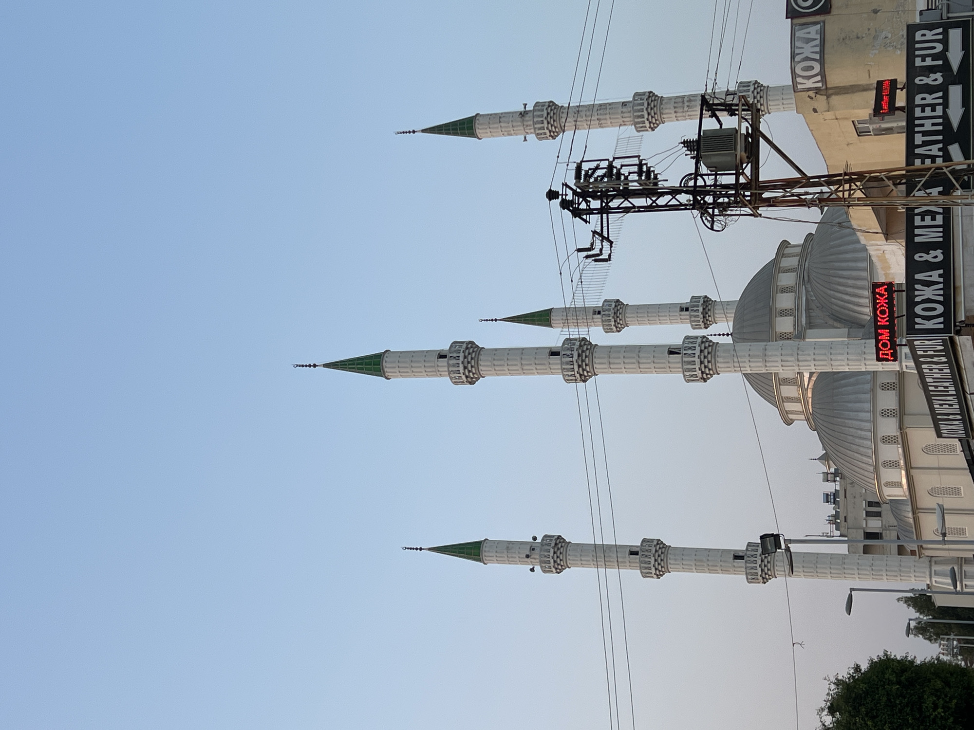 A mosque with several minarets and domes is visible against a clear sky. In the foreground, an electrical transformer and wires are prominent. Signs with text in Russian and English, including "КОЖА", "DOM КОЖА", and "LEATHER & FUR", are visible on buildings to the right of the mosque. Trees are present on the left side of the image. The scene appears to be in an urban setting.