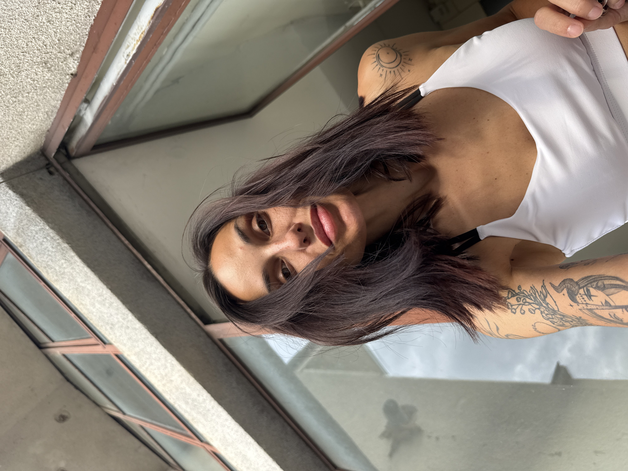 The image captures a young woman looking directly at the camera, with her head tilted and dark brown hair, possibly with subtle reddish or purple undertones, flowing to one side. Strands of her hair partially frame her face, drawing attention to her direct gaze and slightly parted lips. She wears a white top or sports bra with black straps. Visible tattoos include a circular design featuring a crescent moon, sun, and stars on her left shoulder, and a larger, intricate tattoo on her right arm and shoulder, depicting what appears to be a ram's head, wings, and botanical elements.
The setting suggests she is leaning out of a window or positioned in a way that allows a view of a concrete building exterior and the sky. A textured, light gray concrete facade is visible on the left side of the frame, alongside a weathered brown window frame. Through the window glass, a reflection of an overcast or cloudy sky can be seen, indicating it is daytime. The architectural elements and general appearance are consistent with an urban environment in Pleven, Bulgaria. No visible text is present.