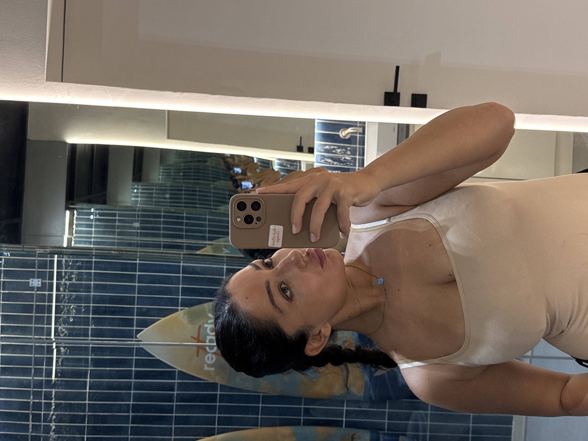 A woman is taking a selfie in a bathroom. She is holding a tan-colored iPhone with three rear-facing lenses. She is wearing a light beige or tan form-fitting bodysuit or leotard and a delicate necklace with a small blue charm. Her dark hair is braided. The bathroom features blue rectangular tiles and a mirrored wall behind her, reflecting the tiling. A surfboard with the text "Ready" is visible in the reflection. The bathroom appears to be well-lit with white ambient light and recessed lighting above the mirror. The location is Kuta Selatan, Indonesia.