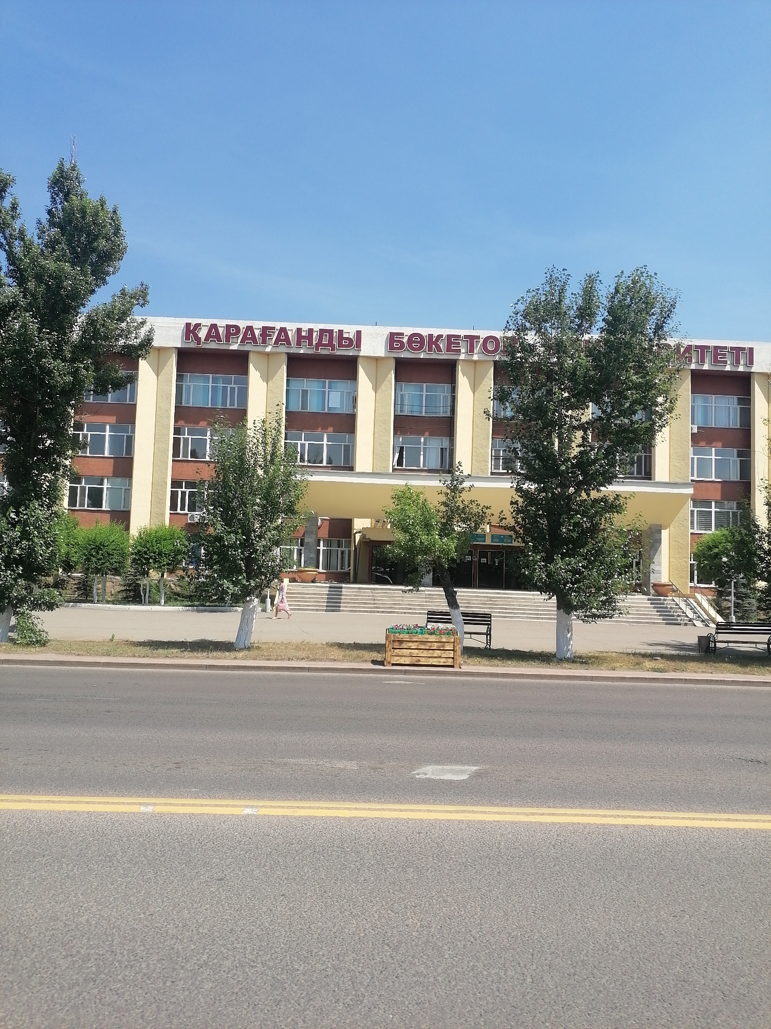 The image captures the facade of a yellow and beige building with maroon lettering that reads "ҚАРАҒАНДЫ БӨКЕТОВ УНИВЕРСИТЕТІ" (Karaganda Buketov University). The building has multiple floors with numerous windows. In front of the building, there are steps leading up to the entrance, and some trees and bushes are visible. To the left of the steps, there is a wooden planter box with flowers. Two park benches are also present. A person in a light-colored dress is walking up the steps on the left side of the entrance. A road with yellow double lines runs across the foreground, indicating a street in front of the university. The sky is clear and blue.