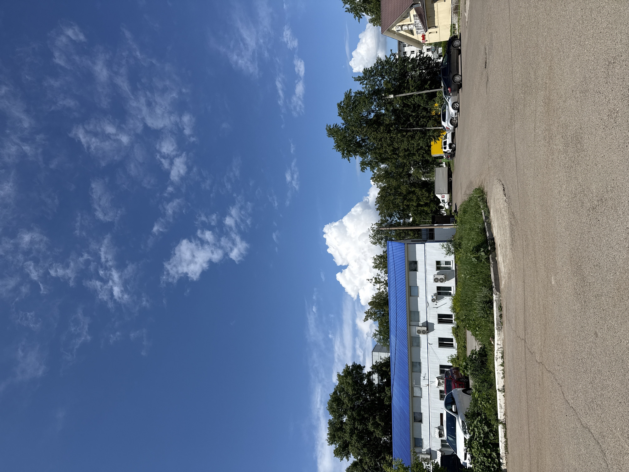 The image depicts a street scene in Chaikovsky, Russia, on a sunny day. A building with a bright blue roof runs along the edge of the street. The building is white with multiple windows and air conditioning units. A few cars are parked in front of the building, obscured by vegetation. The street is asphalt and stretches into the distance, where other buildings and parked cars are visible. Trees are interspersed between the buildings. The sky is blue and filled with scattered white clouds.