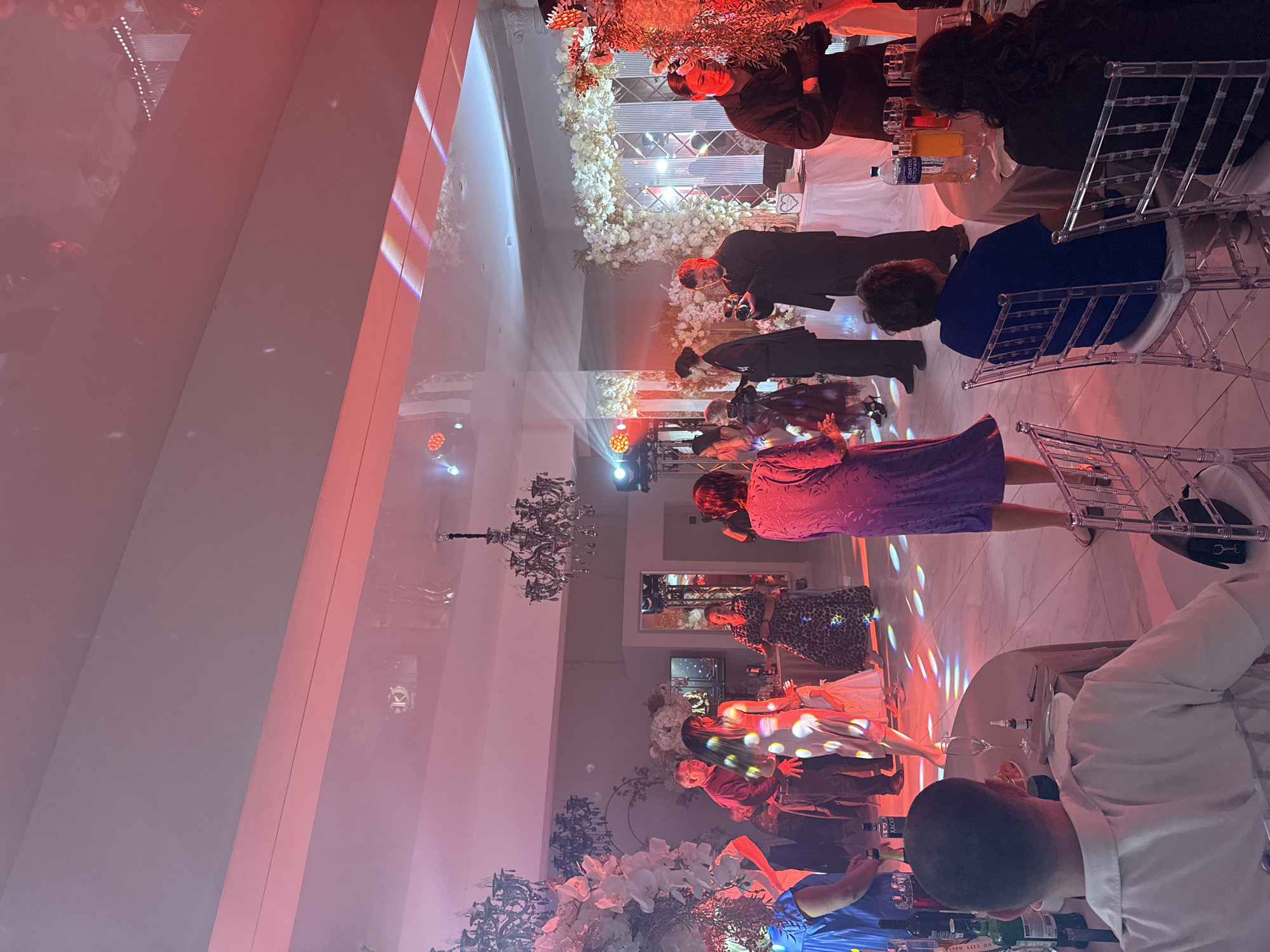 The image depicts a wedding reception in Perm, Russia, with approximately twenty people visible. The room is decorated with floral arrangements and chandeliers, and the lighting is predominantly red and pink, with colorful light projections on the floor. Several individuals are dancing in the center of the room, while others are seated at tables or observing the festivities. A man is filming the event with a video camera, and a DJ booth is visible in the background. The overall atmosphere appears celebratory.