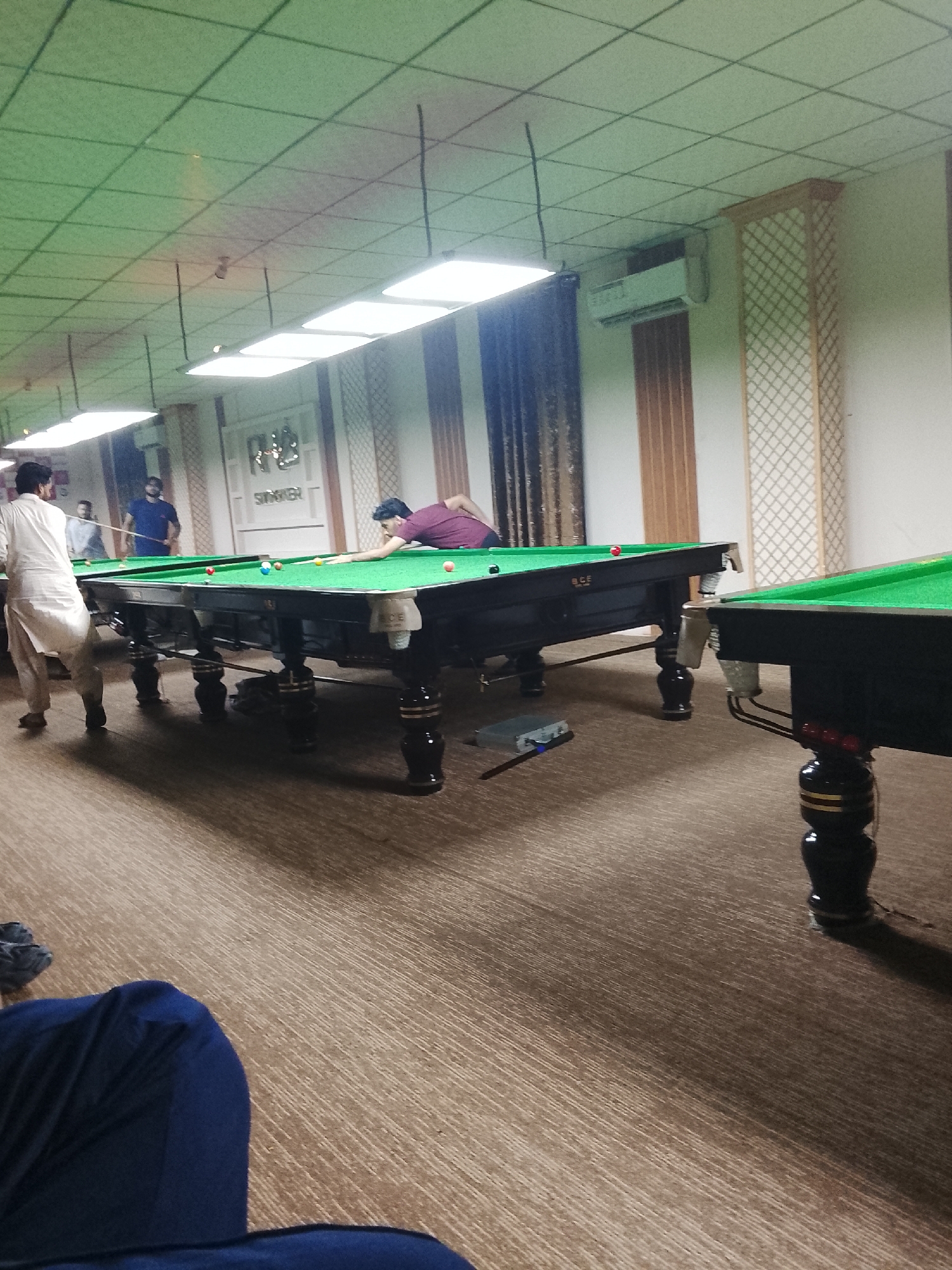The image captures an indoor scene within a snooker or billiards hall, showing several men engaged in games. In the foreground, the back of a person wearing a dark blue shirt is visible, seated or leaning low.
The main action centers around two snooker tables with green felt surfaces and dark, ornately carved wooden legs. On the left table, a man in a white, traditional long shirt (kurta) stands with his back to the viewer, appearing to observe the game or waiting for his turn. Further back at the same table, another man in a maroon shirt is bent over, taking a shot with a cue stick. Snooker balls are scattered on the table. Another man in a blue shirt is visible standing behind the first table. To the right, part of a second snooker table is visible, also with green felt and dark legs.
The setting appears to be a dedicated playing area. The ceiling is made of light-colored acoustic tiles with several bright, rectangular fluorescent light fixtures providing ample illumination. The walls are paneled, featuring both plain sections and sections with a decorative lattice or grid pattern in light wood tones. An air conditioning unit is mounted high on one of the walls. The floor is covered with a light brown, patterned carpet.
Notable details include a small blue electronic device or box on the carpet near the left snooker table, and a snooker cue rest lying on the floor. The tables are professional-grade, with "BCE" visible on the side of the left table. Above an archway or doorway in the background, the word "SNOOKER" is clearly visible, reinforcing the venue's purpose. The overall atmosphere suggests a casual but focused sporting environment.
