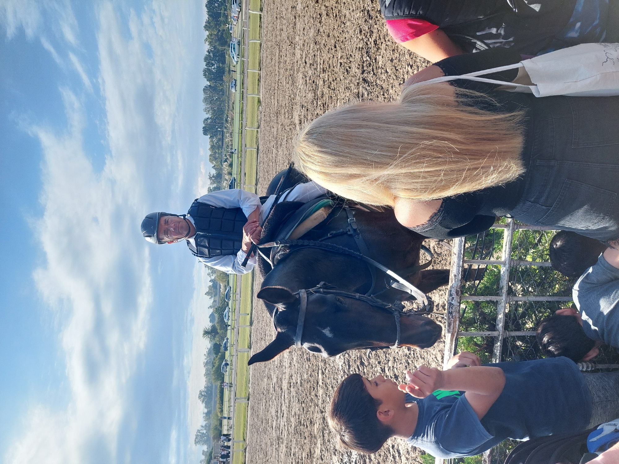 This image captures a lively scene at a horse racing track, likely in Buenos Aires, Argentina, during a sunny day. In the center, a male jockey, wearing a grey helmet, a light-colored long-sleeved shirt, and a dark protective vest, is mounted on a dark brown horse. He looks towards the viewer with a friendly smile, holding the reins. The horse is saddled and bridled, standing calmly.
To the right, a blonde-haired woman is seen from behind, wearing a black top and dark jeans, with a white tote bag on her shoulder. She is positioned near the horse's head, observing the interaction. Below her, separated by a white metal fence, two boys are interacting with the horse. The boy in the foreground, wearing a blue t-shirt with a green emblem, gazes up at the horse with an expression of curiosity or excitement, his hands clasped together. Another boy is partially visible behind him.
The background reveals the expanse of the dirt racing track, a green grassy area beyond it, and a line of trees with parked cars further in the distance. The sky is bright with scattered white clouds, indicating clear weather. The scene depicts a moment of connection between a jockey and his horse with spectators, possibly during a public event, meet-and-greet, or training session at the track. There is no discernible text visible in the image.