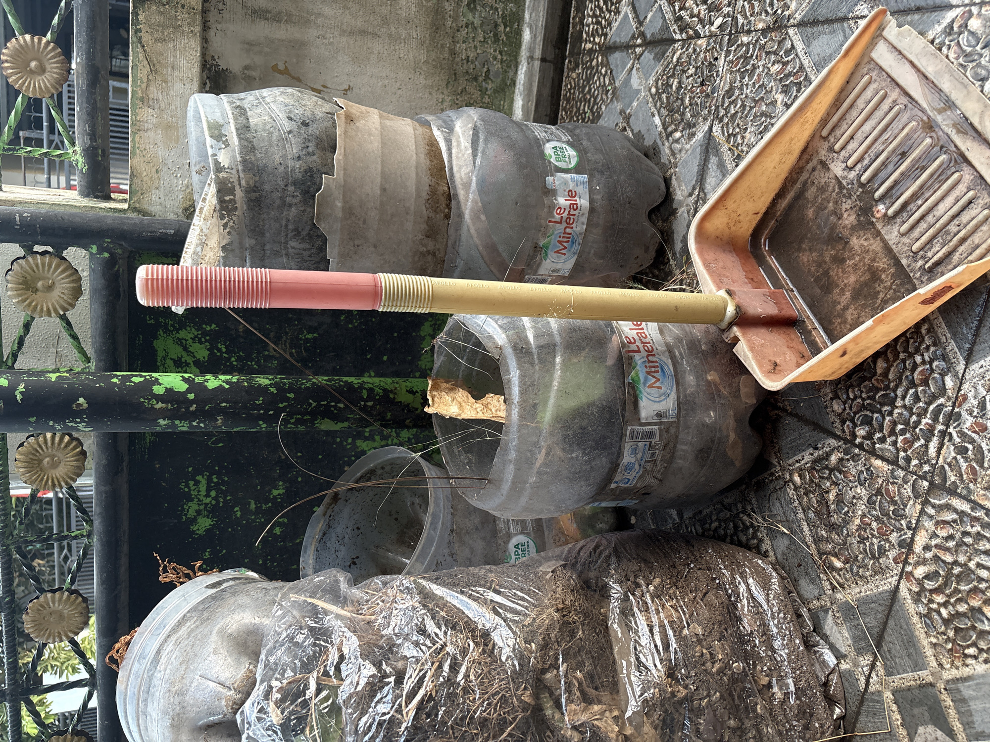 The image shows a collection of repurposed plastic bottles and a dustpan on a tiled patio, likely in Palimanan, Indonesia. In the foreground, a rusty orange dustpan with a yellow handle is prominent, holding some debris and a small amount of liquid. Behind it, a stack of large clear plastic bottles, some cut and used as planters, are visible. One bottle contains dried plant material and a large brown leaf. Another bottle appears to be filled with soil and is covered in plastic sheeting. A tall, pink and yellow handle of a broom or tool is positioned between the bottles. The background features a black metal fence with decorative gold elements and green paint splatters on a post. The ground is paved with grey and beige stone tiles, and there are some dry leaves and twigs scattered around. The scene suggests a makeshift gardening setup or storage area, possibly in a residential or backyard setting.