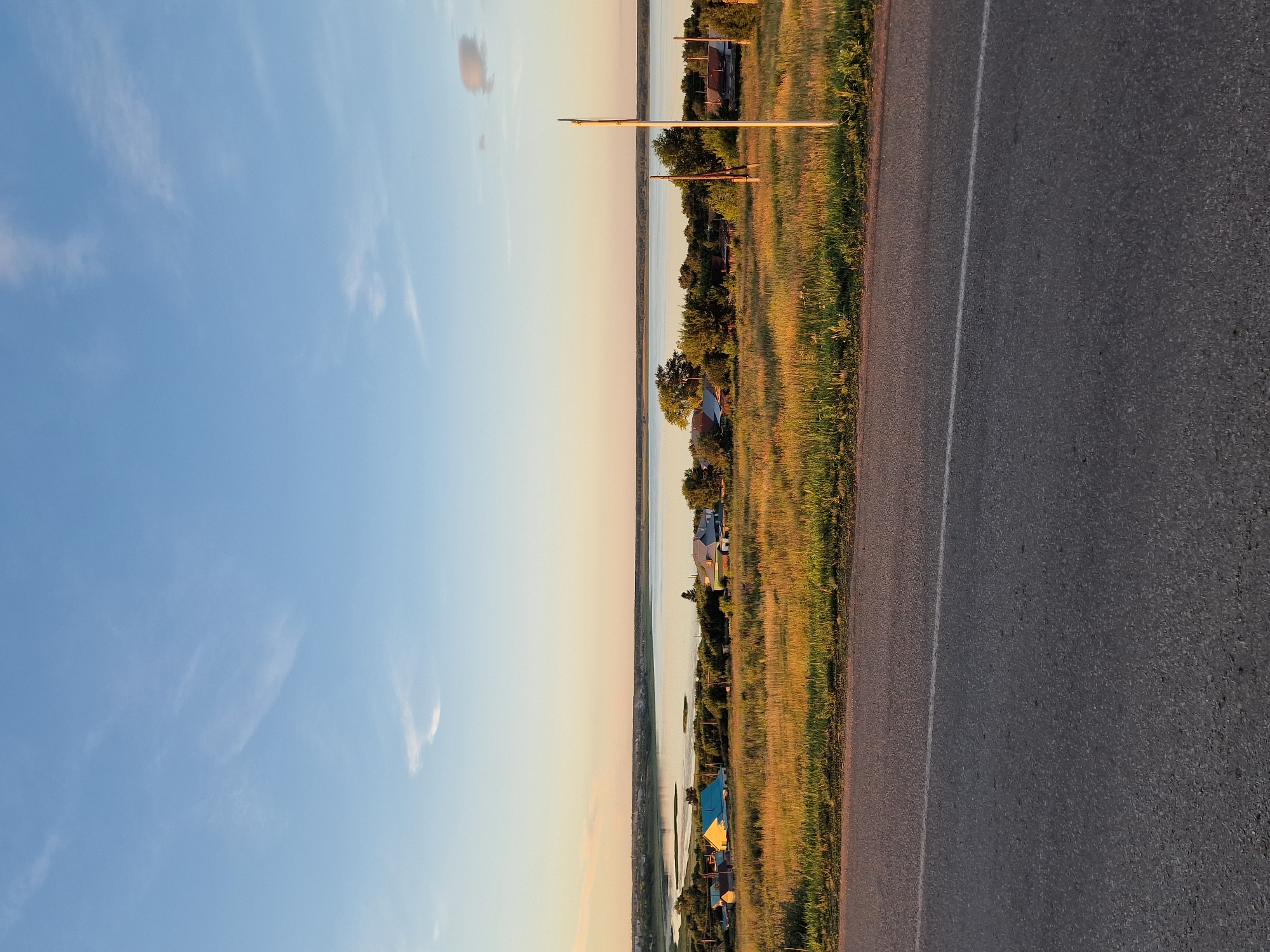 The image depicts a rural landscape under a clear blue sky with wispy white clouds. In the foreground, a grey asphalt road with a white line marking the lane is visible. Beyond the road, a grassy field slopes gently upwards, dotted with wildflowers and tall grasses illuminated by the warm glow of what appears to be sunset. Several houses with pitched roofs are scattered across the field, some with brightly colored roofs. In the middle ground, a tranquil body of water, possibly a lake or river, stretches across the horizon, reflecting the soft light of the sky. A line of trees and vegetation borders the water. A thin, white pole stands vertically on the right side of the image in the grassy field. The overall scene suggests a peaceful, natural environment in a rural area. There are no people or animals visible in the image. The location is not Kashgar City, China; it is a rural landscape.