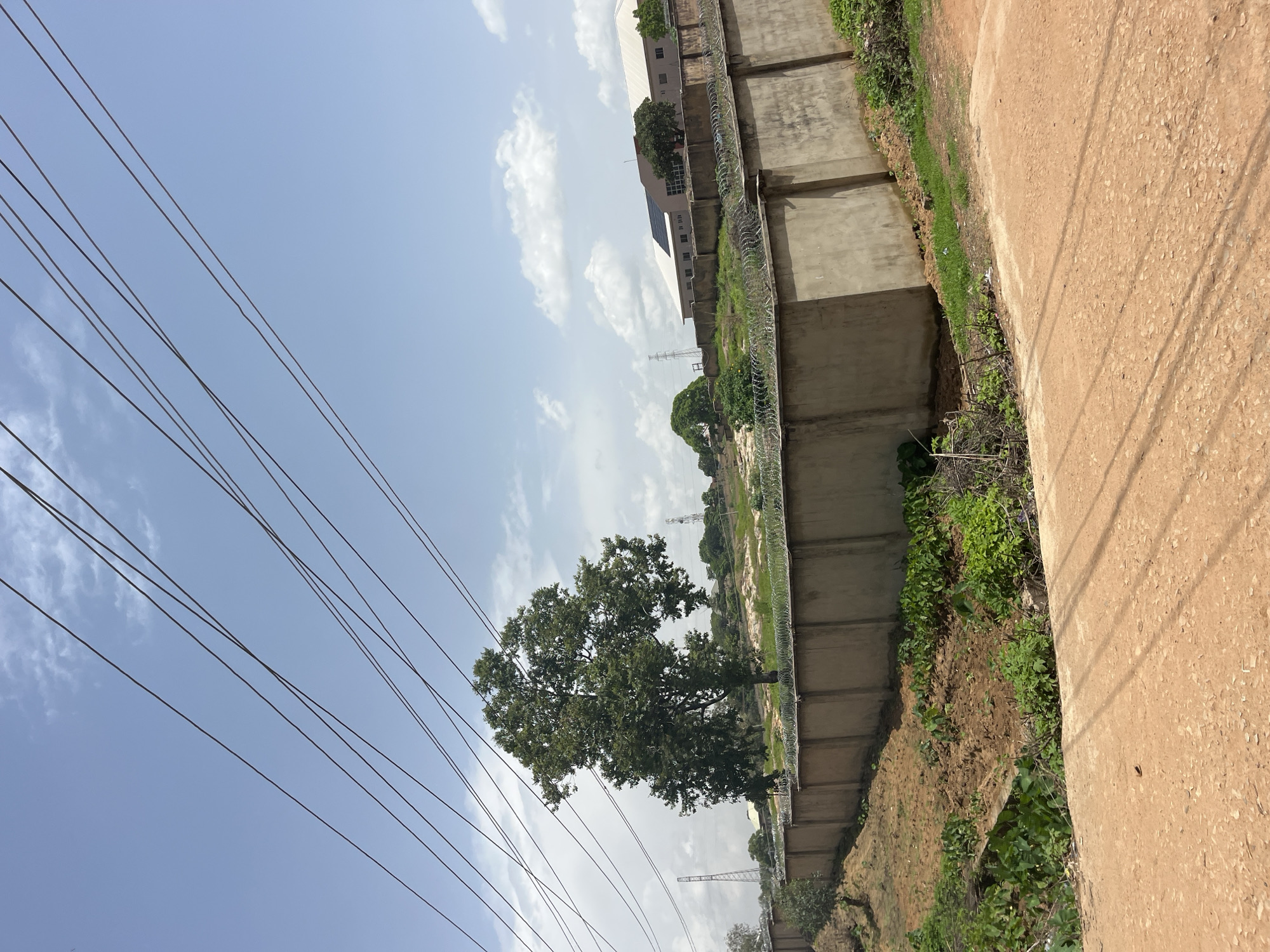 The image depicts a view of a concrete wall topped with barbed wire, likely serving as a perimeter for a property. A large, leafy tree stands prominently behind the wall, casting a shadow on the ground. Beyond the tree, the landscape includes scattered vegetation and what appear to be communication towers in the distance. A building with a light-colored roof is visible further back, suggesting an urban or semi-urban setting. Overhead, several power lines are strung across the sky, which is partly cloudy. The foreground shows a dirt road. The scene appears to be located in Gura Top, Nigeria, given the context of the request.