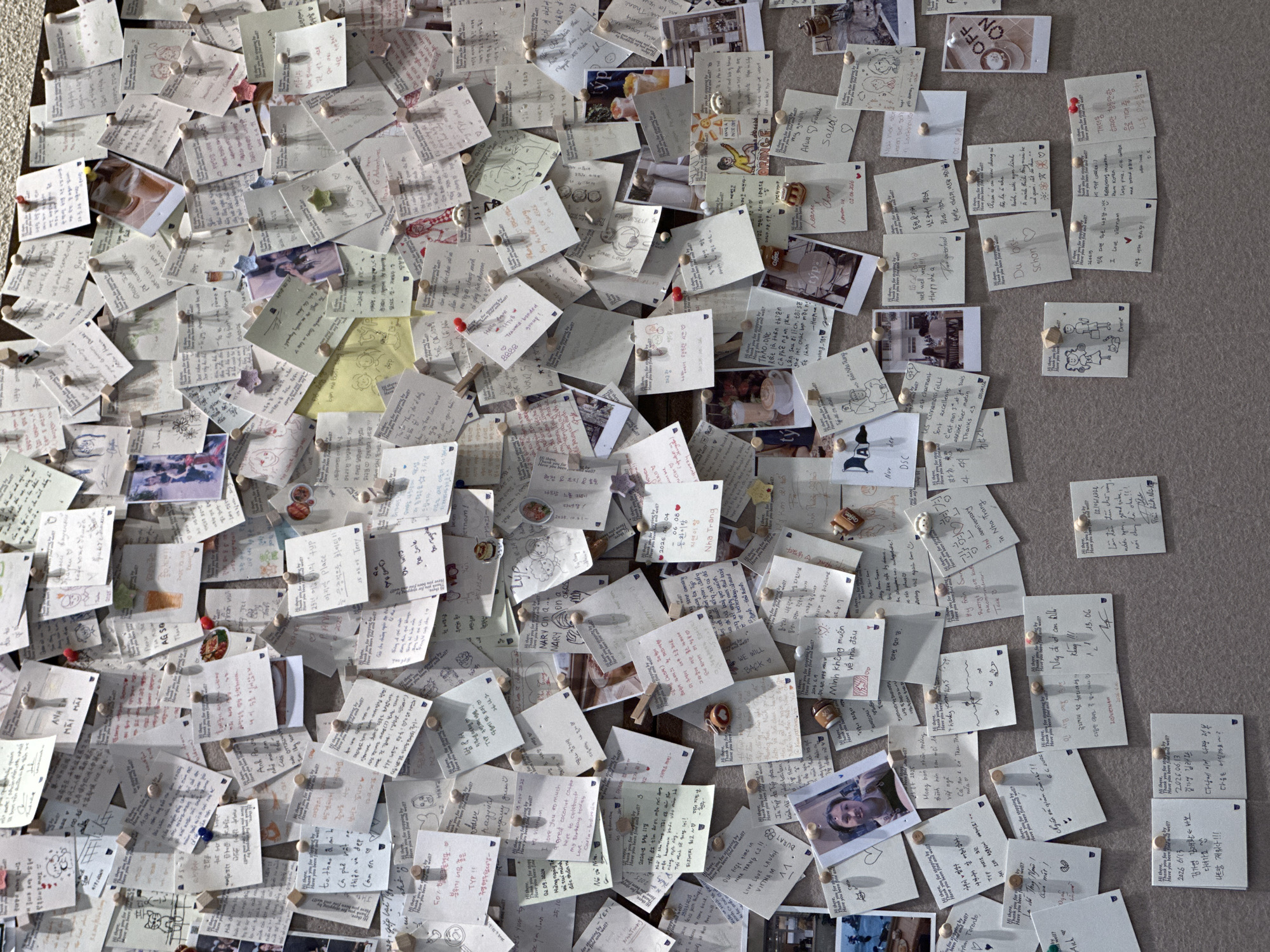 The image displays a close-up, high-angle view of a wall covered in numerous handwritten notes, small photographs, and drawings, pinned up with thumbtacks. The notes are of various sizes and colors, predominantly white, with some yellow and pastel colored papers. Many notes contain text in different languages, including English, Korean, and Vietnamese. Common phrases like "Hi there, Thank you for stopping by, Have you been find and well?" are repeated on multiple notes. Some notes feature drawings of people, animals, and abstract designs. A few small photographs are interspersed among the notes, showing portraits of individuals, scenes of people dining, and beverages. Small decorative items, such as miniature coffee cups and star-shaped pins, are also attached to some notes. The overall presentation suggests a public display of messages, possibly in a cafe, hostel, or tourist information center in Nha Trang Ward, Vietnam. The texture of the wall appears to be a neutral, slightly rough surface. The lighting is even and diffused, indicating an indoor setting.