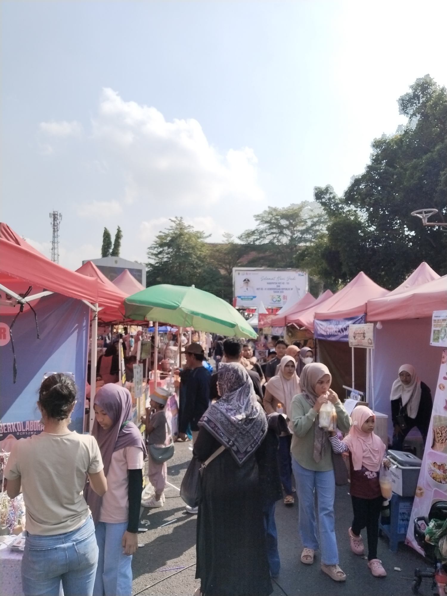The image captures an outdoor market scene, likely in Kecamatan Pati, Indonesia, with several rows of tents providing shade. The sky is bright and partly cloudy, with visible trees and a distant telecommunications tower. A variety of people are browsing and walking through the market, many of whom are wearing headscarves. Some individuals are interacting with vendors or purchasing items. The ground appears to be paved. In the background, there are signs and advertisements. The overall atmosphere suggests a busy and active community event.