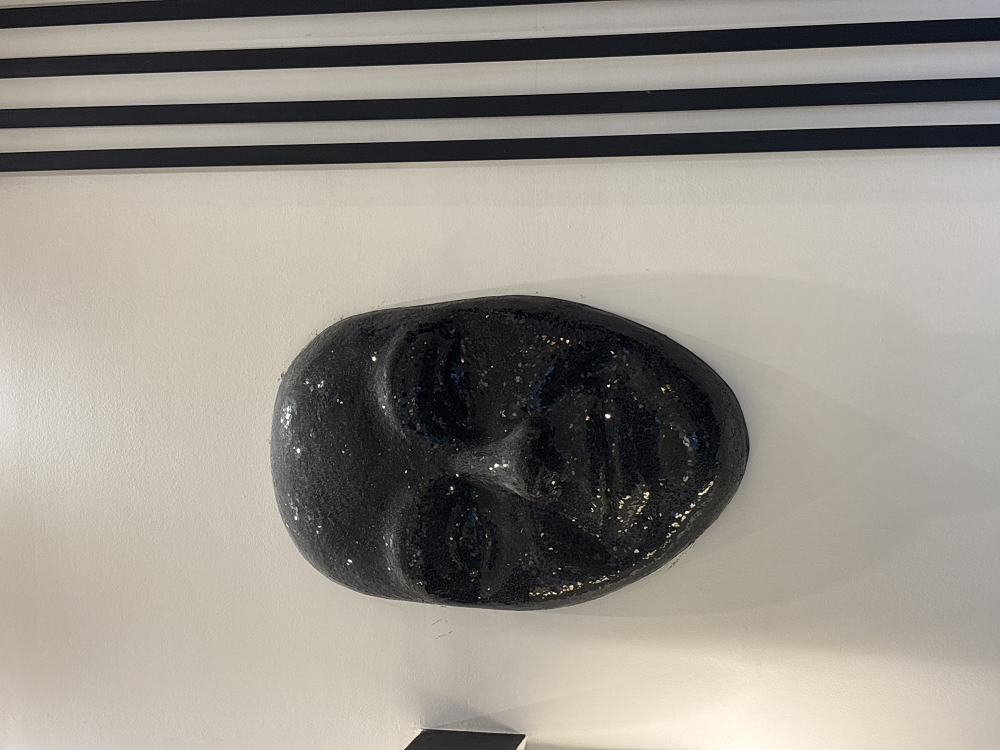 A black, sequined mask is mounted on a white wall. The mask is a human face with visible eyes, nose, and mouth. To the right of the mask, there are black and white vertical stripes on the wall. The background is a plain white wall. The location is identified as a room in the city of Khirbet Al Souk, Jordan.