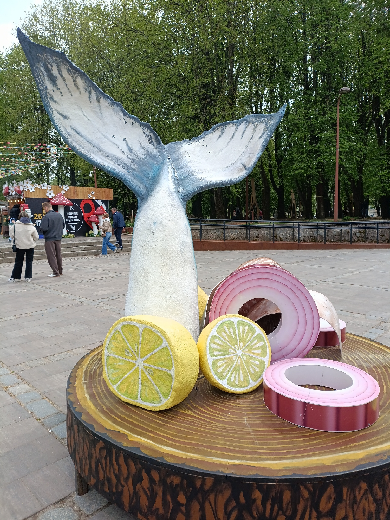 In a public square in Kaliningrad, Russia, a whimsical sculpture rests on a faux tree stump. The sculpture features a large whale tail emerging from the top, surrounded by oversized slices of lemon and red onion. People stroll in the background amidst greenery and buildings.