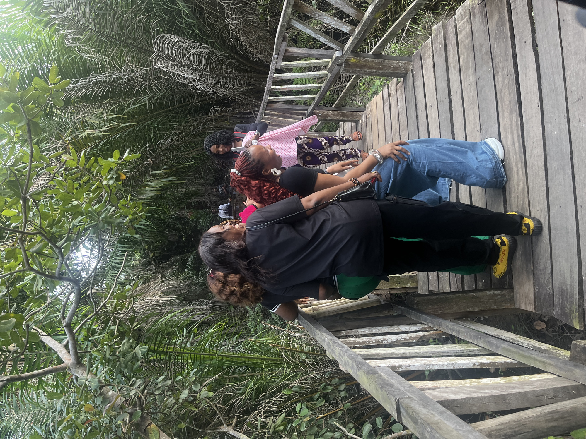 The image depicts a group of women on a wooden boardwalk in a lush, tropical environment, likely in Lekki, Nigeria. The boardwalk, constructed of weathered planks and sturdy wooden railings, winds through dense vegetation dominated by palm trees and broad-leafed plants. Sunlight filters through the canopy, creating dappled lighting.
In the foreground, three women are visible. The woman on the left, facing away from the camera, wears a black t-shirt, green pants, and yellow shoes. She is leaning on the railing, engaged in conversation with the woman next to her. This second woman, also facing away, has dark, curly hair and wears a black t-shirt and black pants. She appears to be looking at something off-camera. The third woman, in the center and slightly further back, has vibrant red curly hair tied up and wears a black t-shirt with wide-leg blue jeans. She is looking down and laughing, her right hand resting on her knee, adorned with a silver bracelet.
Further down the boardwalk, another woman is partially visible, wearing a pink and white polka-dot top and patterned pants. She is holding a pink cloth. Behind her, another woman in a black jacket and patterned pants is visible. The overall impression is of a casual outing or exploration in a natural setting.