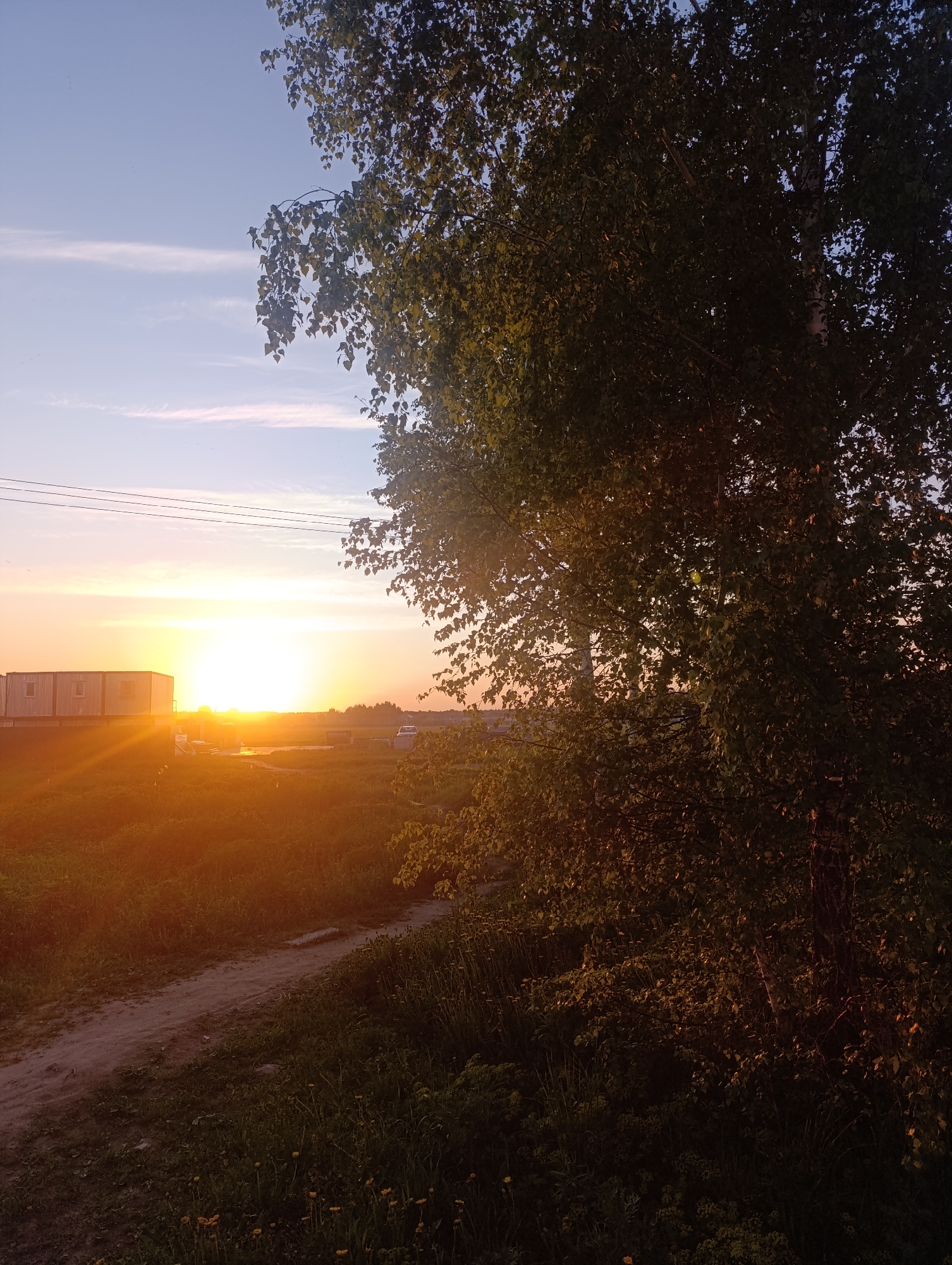 The image depicts a sunset scene. In the distance, there's a structure that appears to be a building or trailer. The sun is positioned on the horizon, radiating a bright orange and yellow glow. In the foreground, there's a tree with dense foliage. A dirt path is visible in the lower portion of the image. The location is Unknown, Unknown.