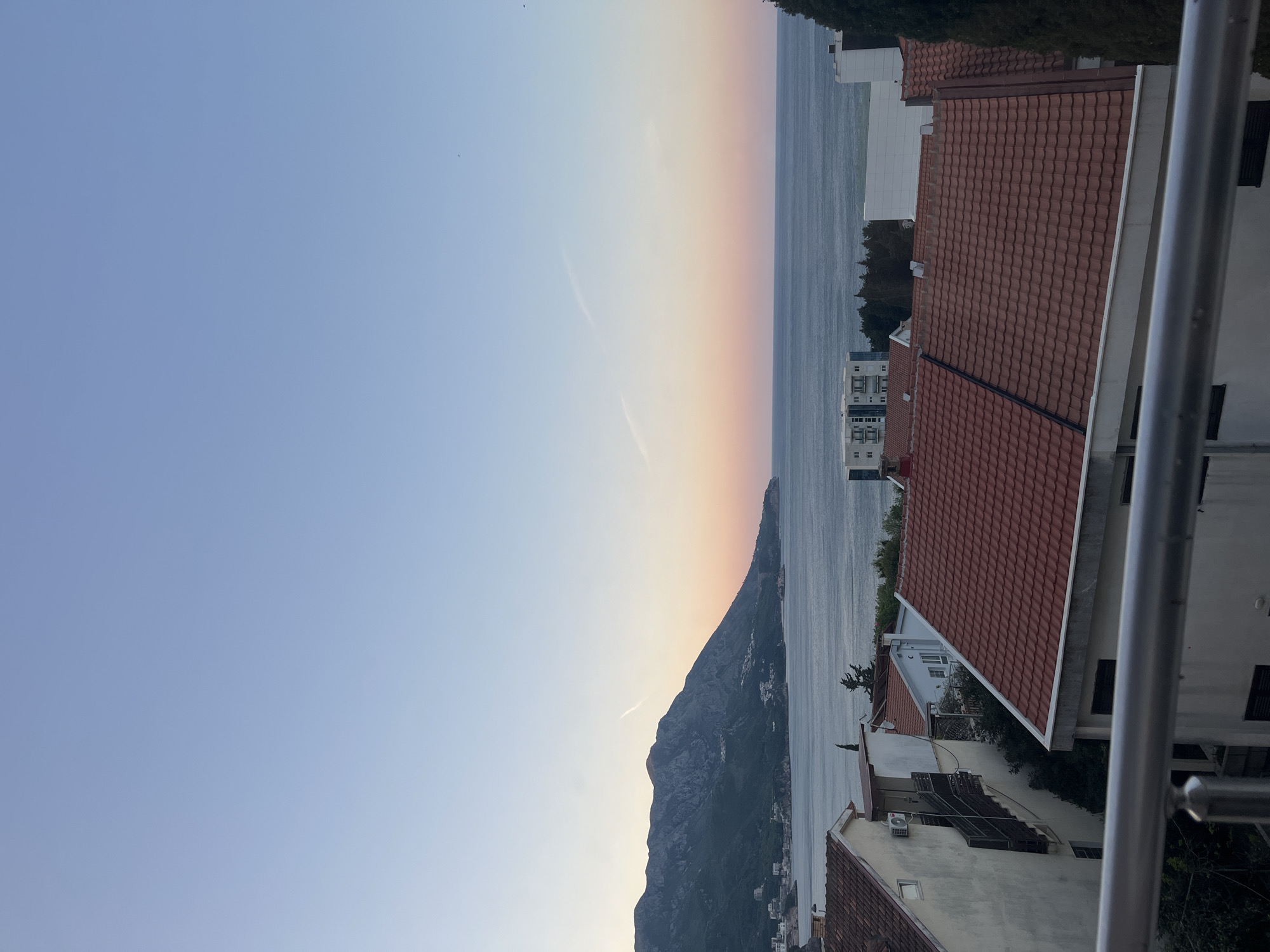 The image depicts a coastal scene with a mountain range along the sea. The sky transitions from blue to orange, indicating either sunrise or sunset. Buildings with red tile roofs are visible in the foreground and along the coast. A metallic railing is partially visible, suggesting the photo was taken from a balcony. The location appears to be a populated area along the coastline with natural landscapes.